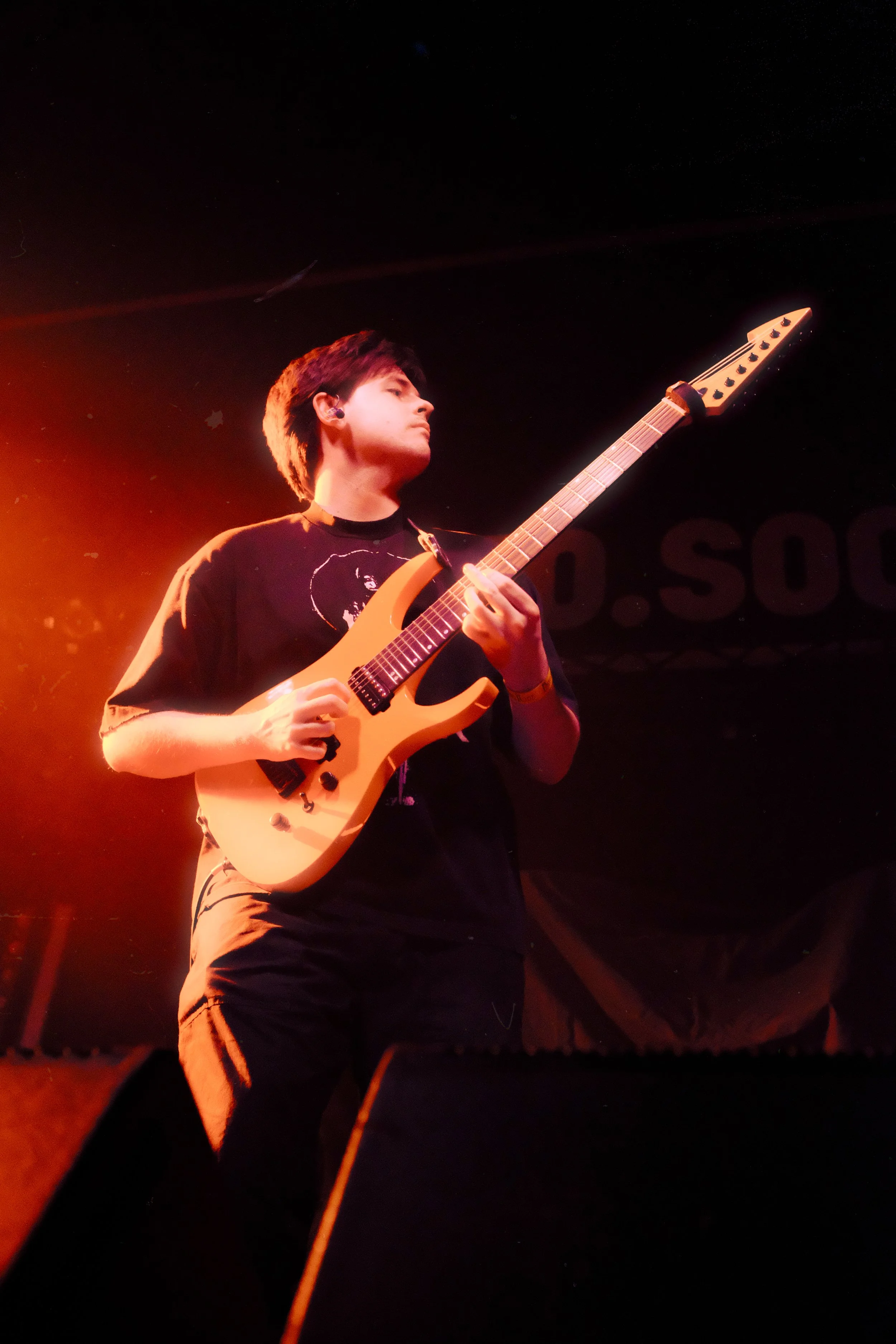A young man playing an orange electric guitar on stage under red lighting.
