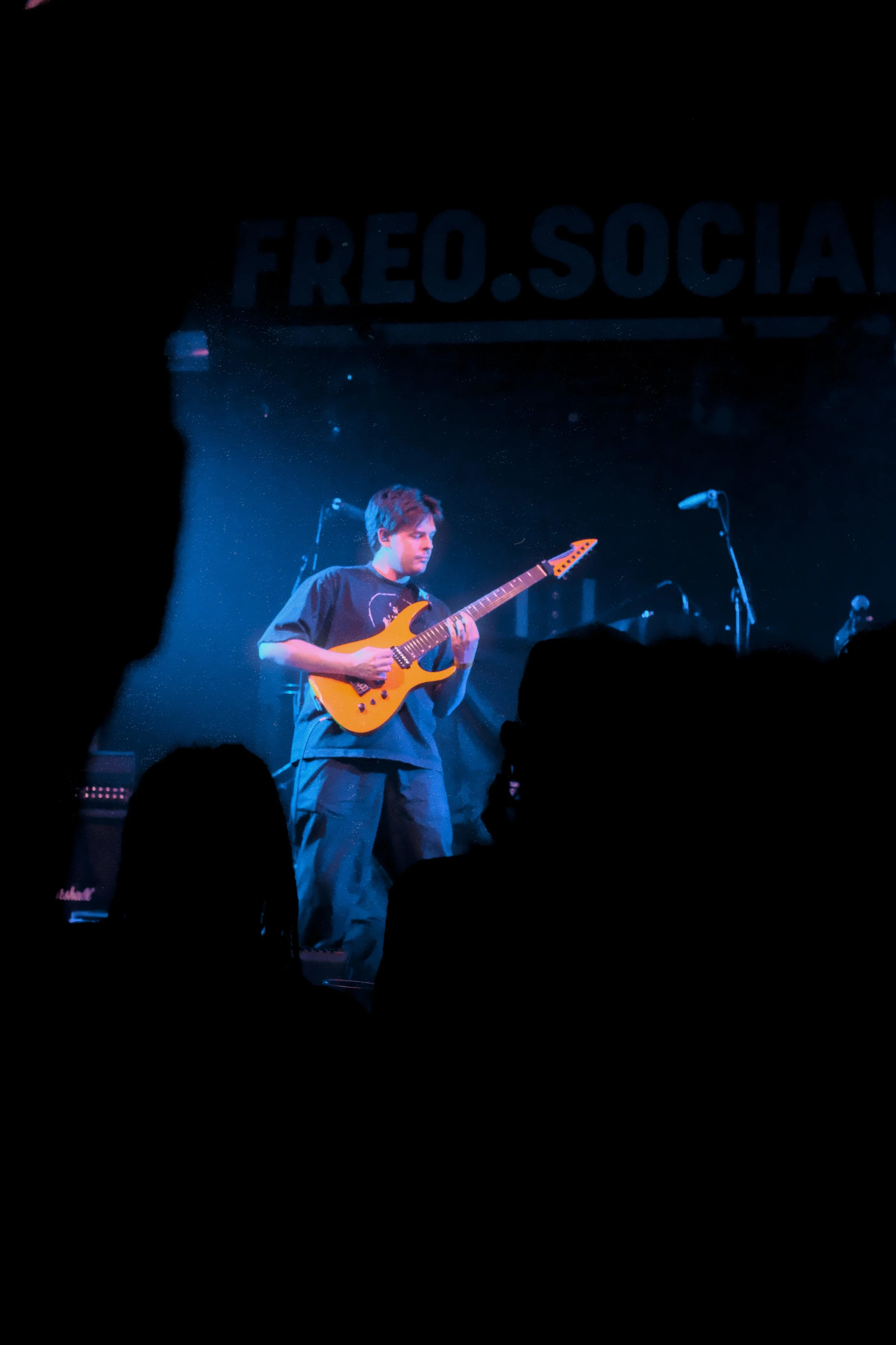 A young man playing an orange electric guitar on stage during a concert with a blue lighting background. There are microphones and speakers visible on stage, and the audience silhouettes in the foreground.