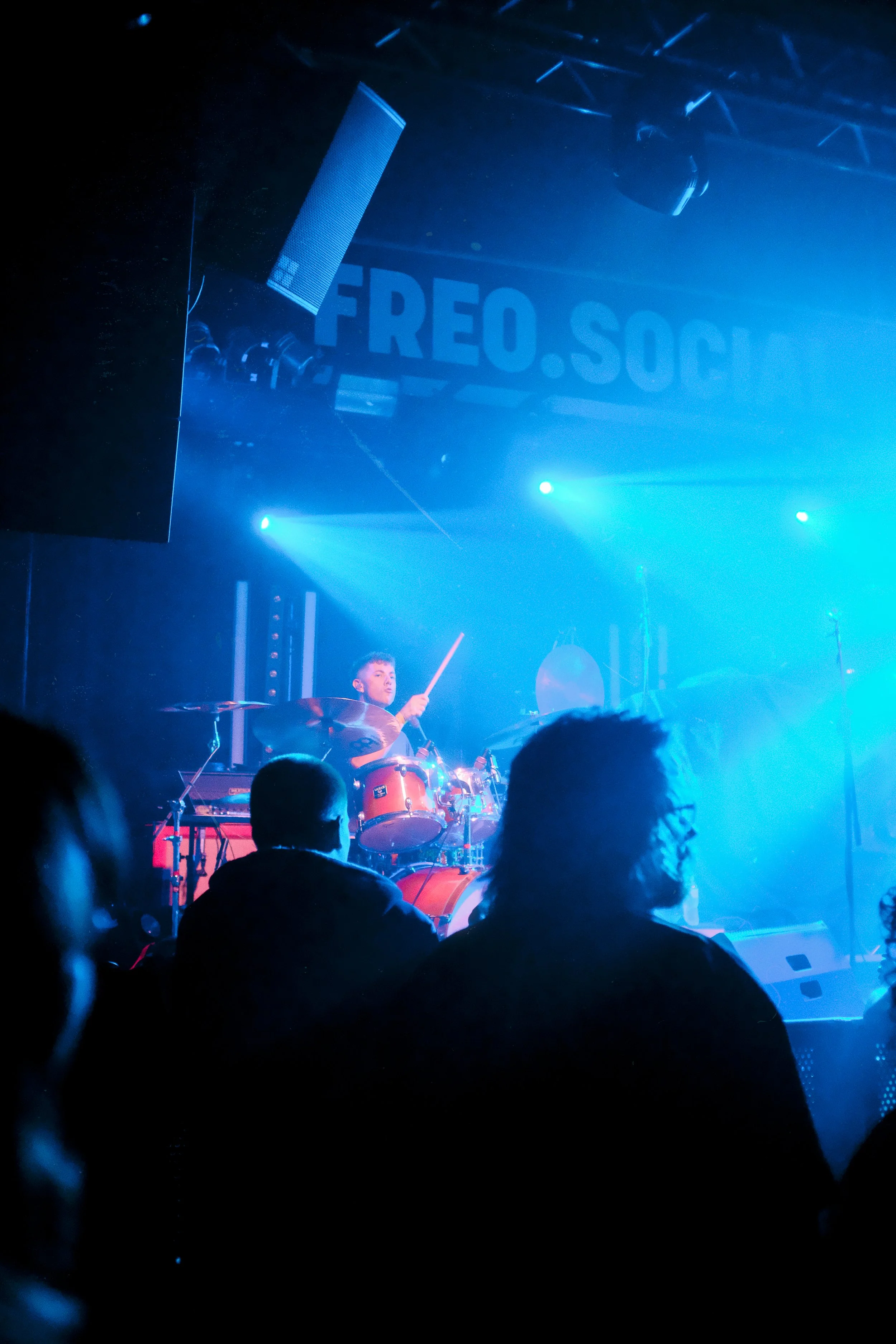 A band performing live on stage with blue lighting, with a drummer playing and a large sign reading 'FREO.SOC' in the background.