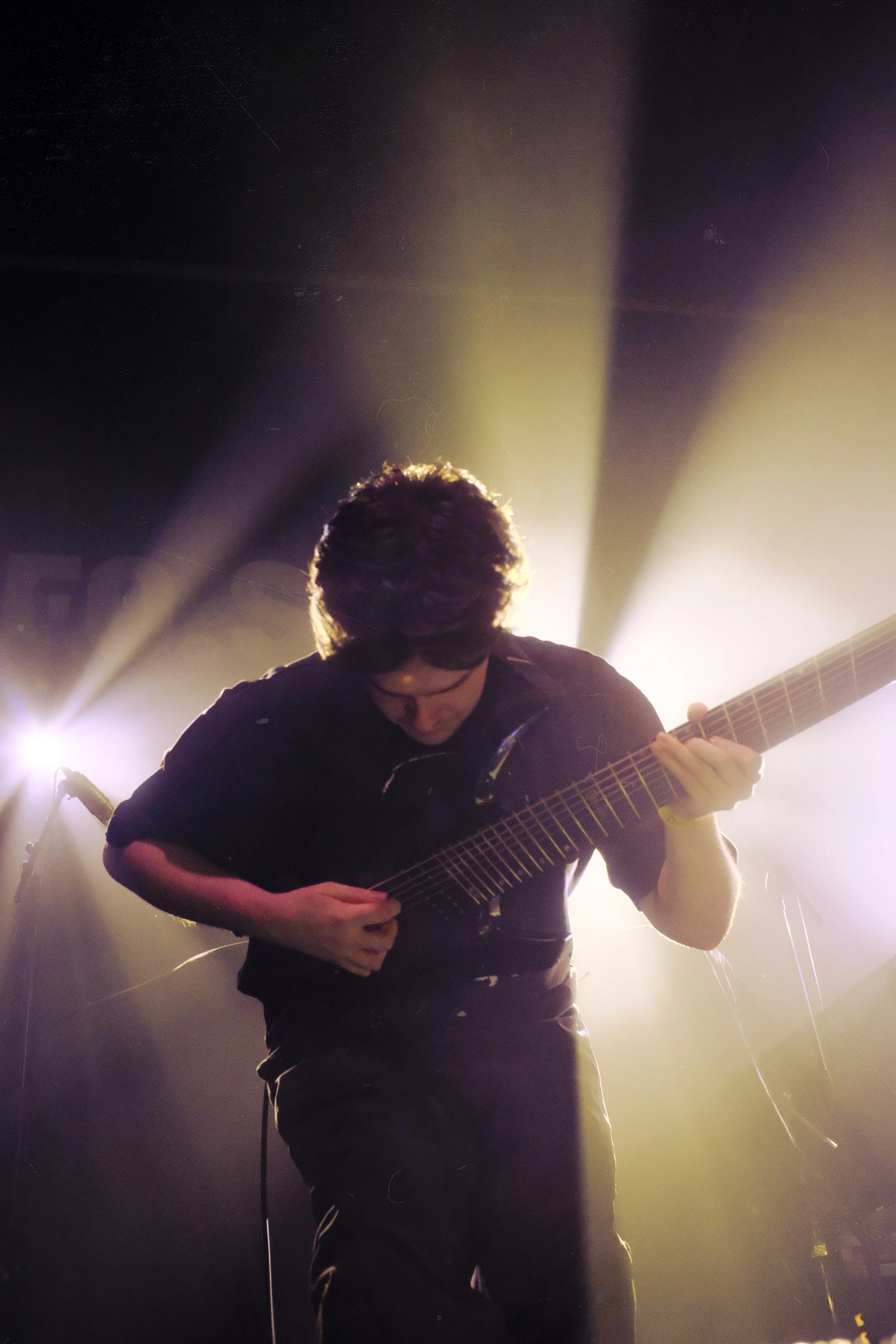 A musician playing a guitar on stage, backlit with bright yellow and white lights creating a halo effect behind him.