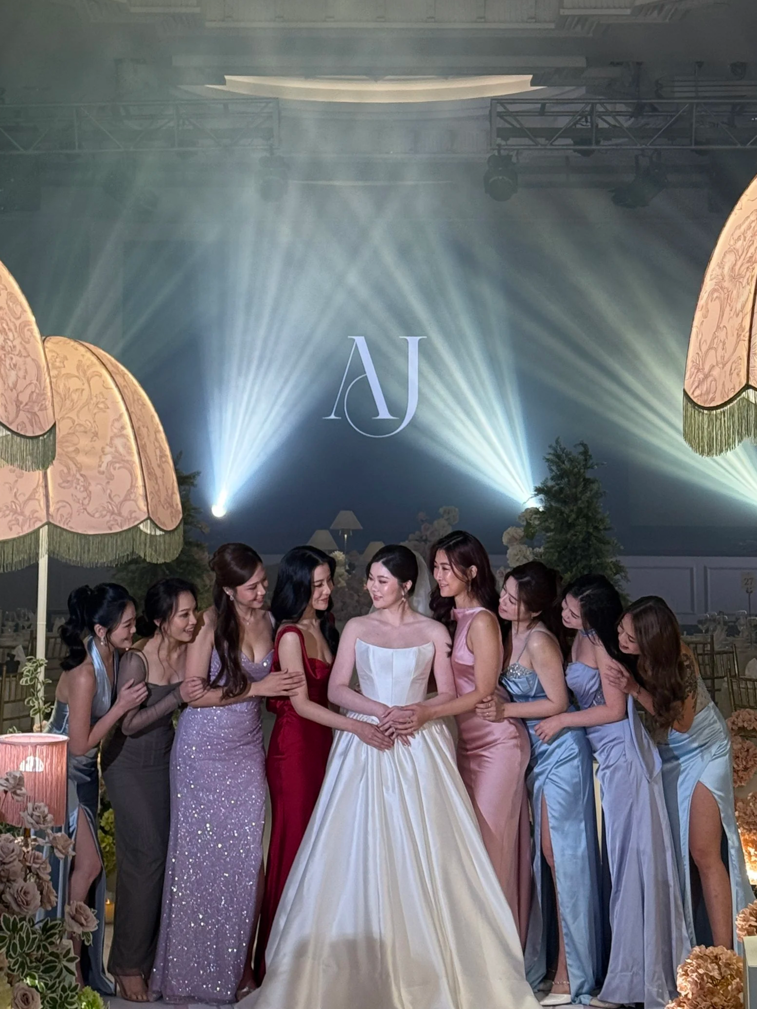 Group of women in colorful evening dresses standing together at a wedding reception with a bride in a white gown at the center, decorated with floral arrangements, lighting, and large pink umbrellas in the background.