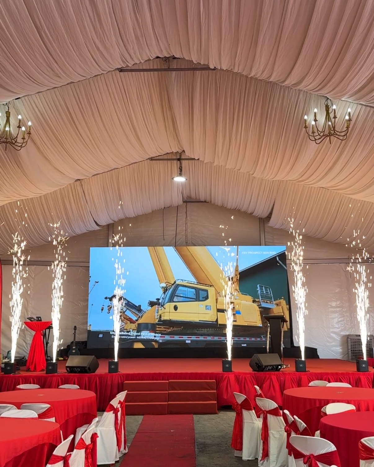 Indoor event space with a stage, large screen displaying construction equipment, red carpet, and tables with red tablecloths and white chairs decorated with red ribbons, pink draped ceiling, chandeliers, and sparklers.