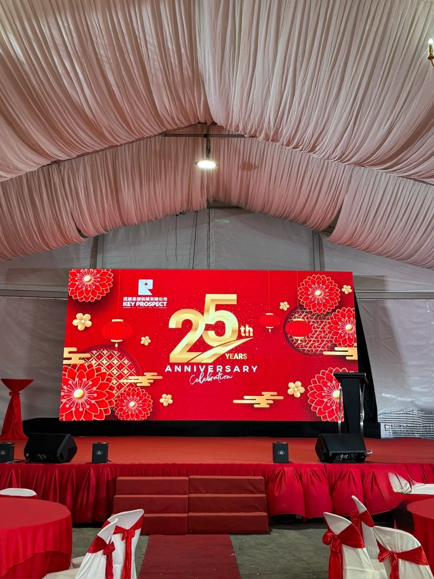 Stage decorated with red and gold for a 25th anniversary celebration, featuring a large digital screen displaying the event name and theme, surrounded by red floral and lantern motifs, with seating arrangements draped in white covers with red ribbons.