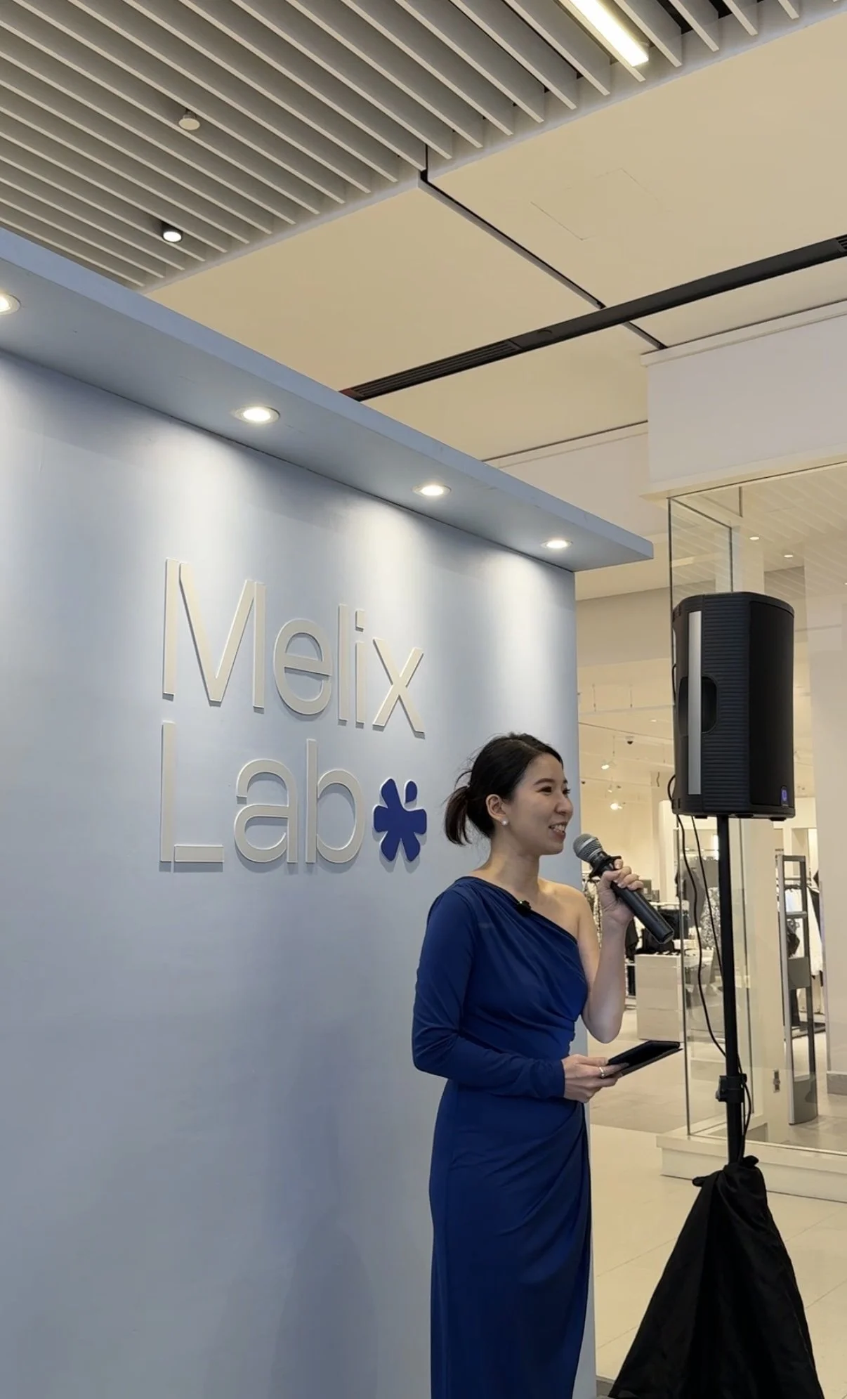 A woman in a blue dress speaking into a microphone at a Melix Lab event inside a modern building.