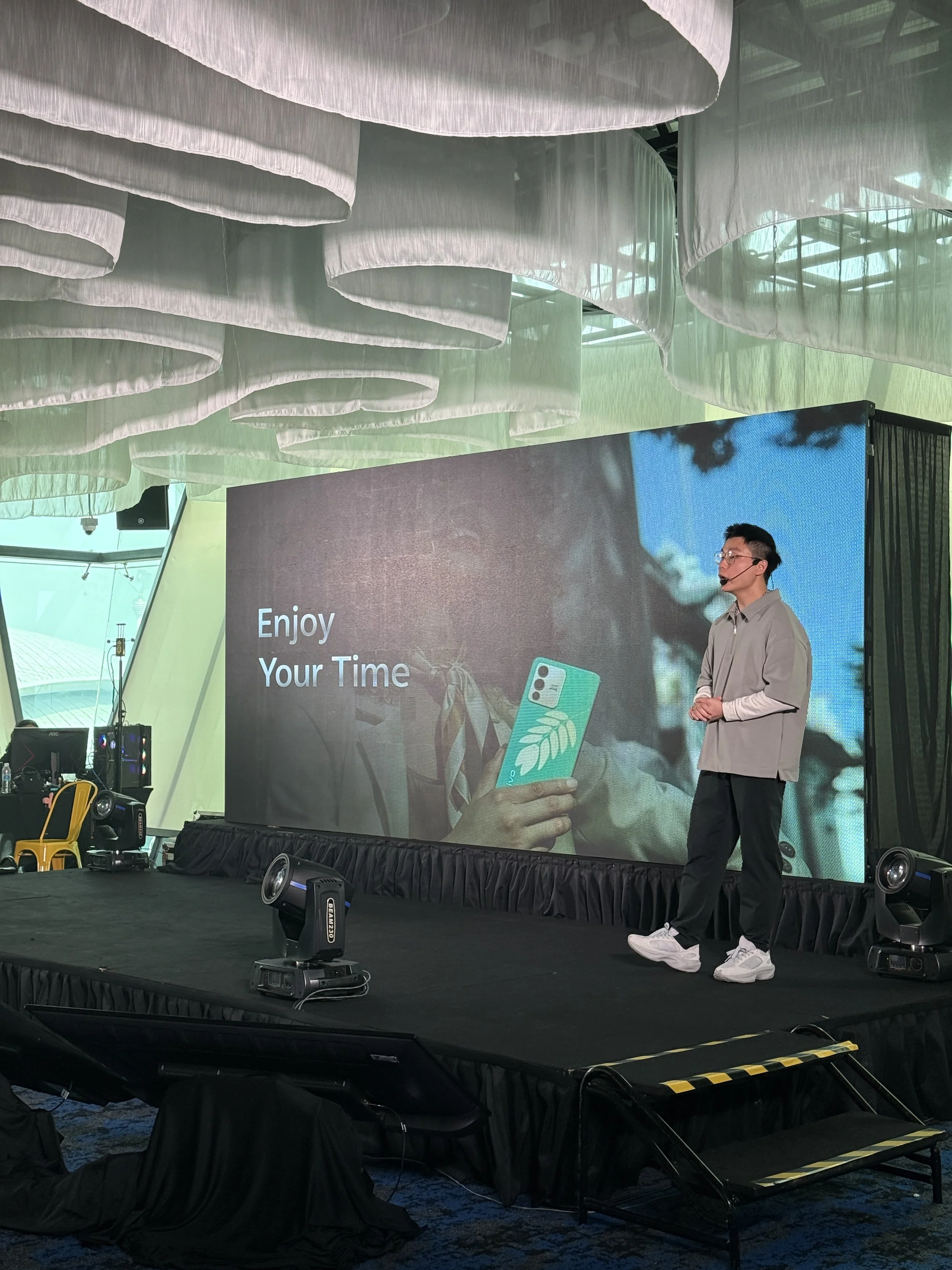 A man standing on a stage in front of a large LED screen, explaining the launch of Vivo's latest phone to the crowd! He is using a Wireless Lapel Microphone