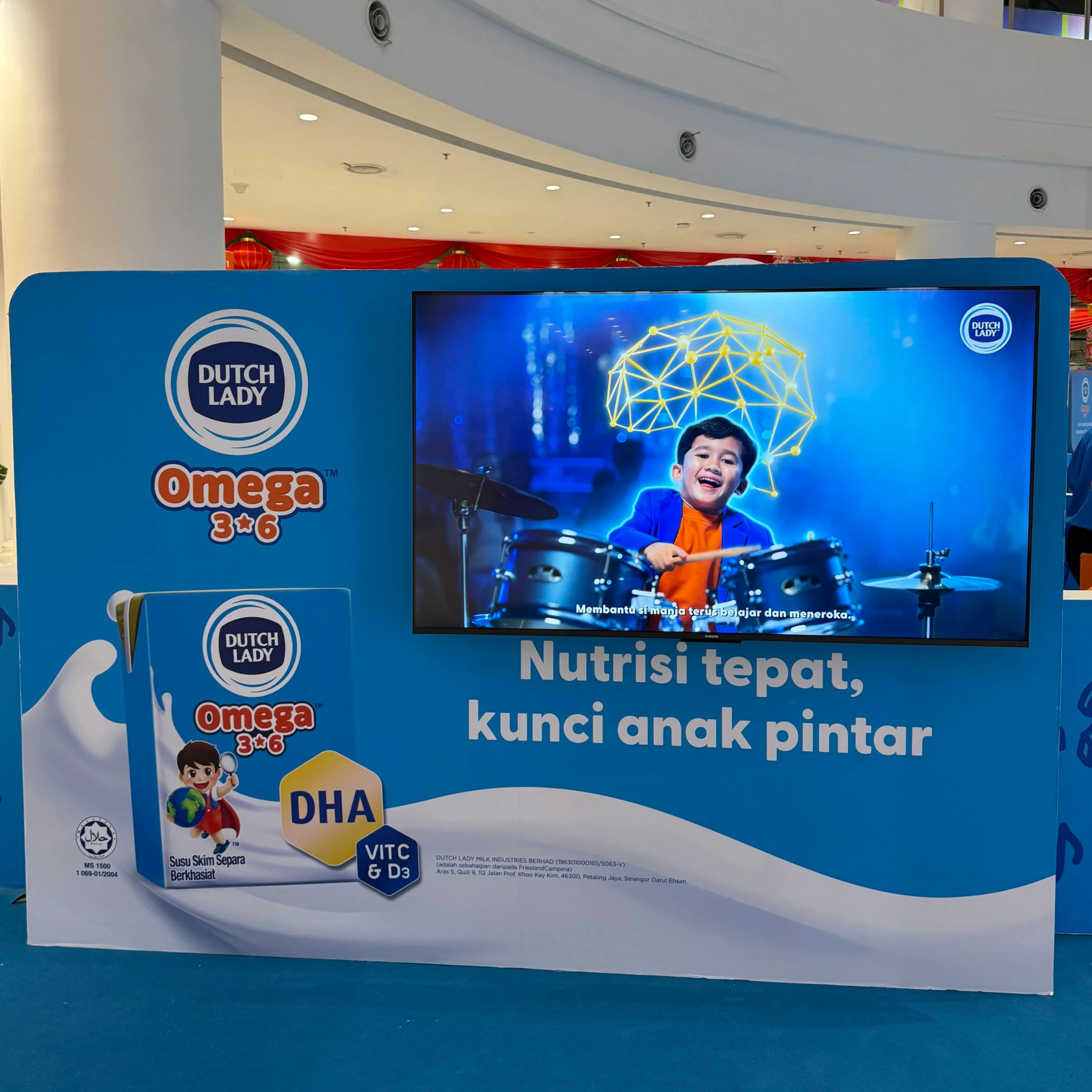 A promotional display for Dutch Lady Omega 3-6 milk featuring a young boy playing drums, with a digital background of a neon brain icon. The text promotes proper nutrition as the key to a smart child, with product branding and nutritional highlights such as DHA, VITC, and D3.