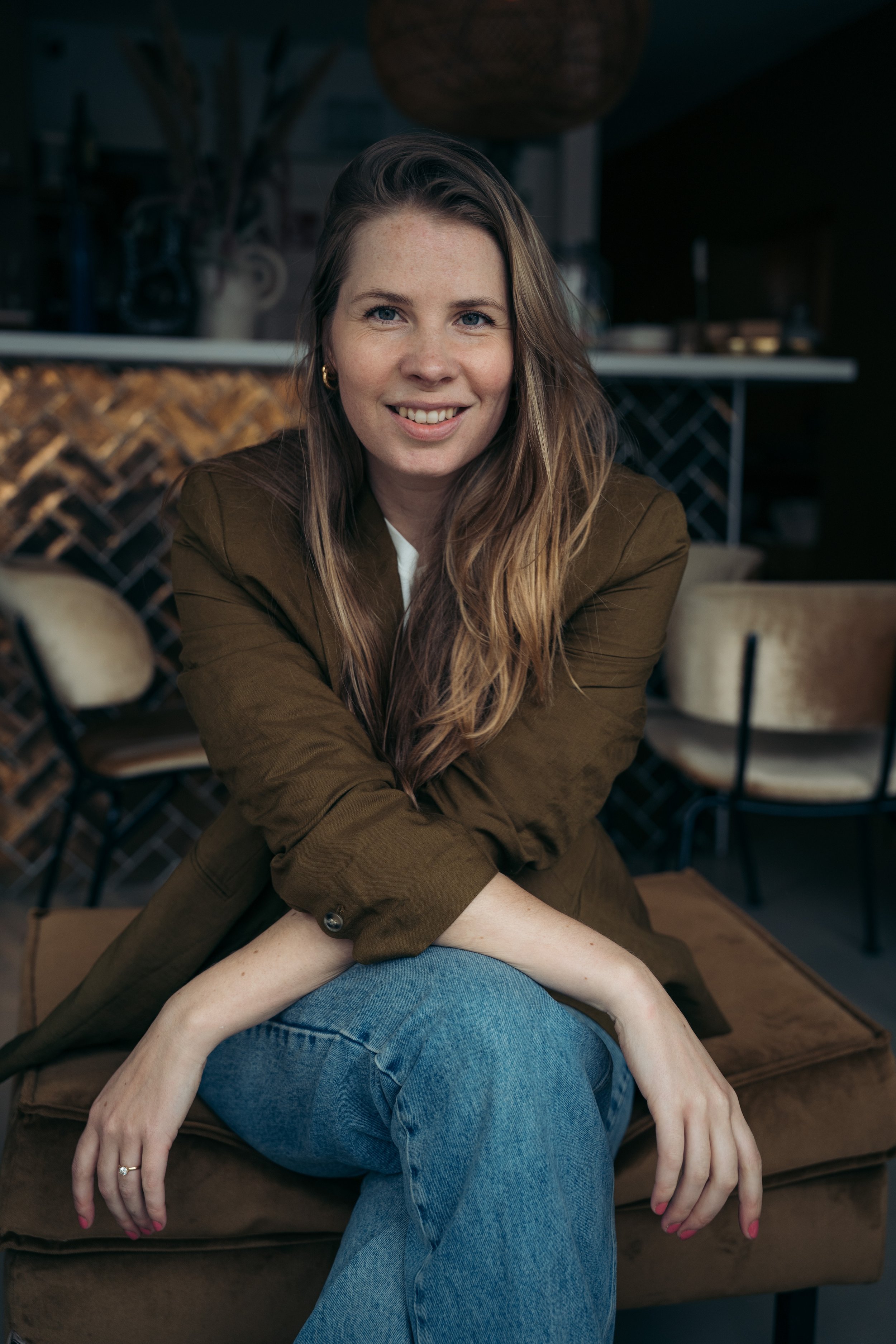 Portret van een lachende vrouw met bruin haar, zit op een bank in een interieur met houten en keramische decoraties op de achtergrond.