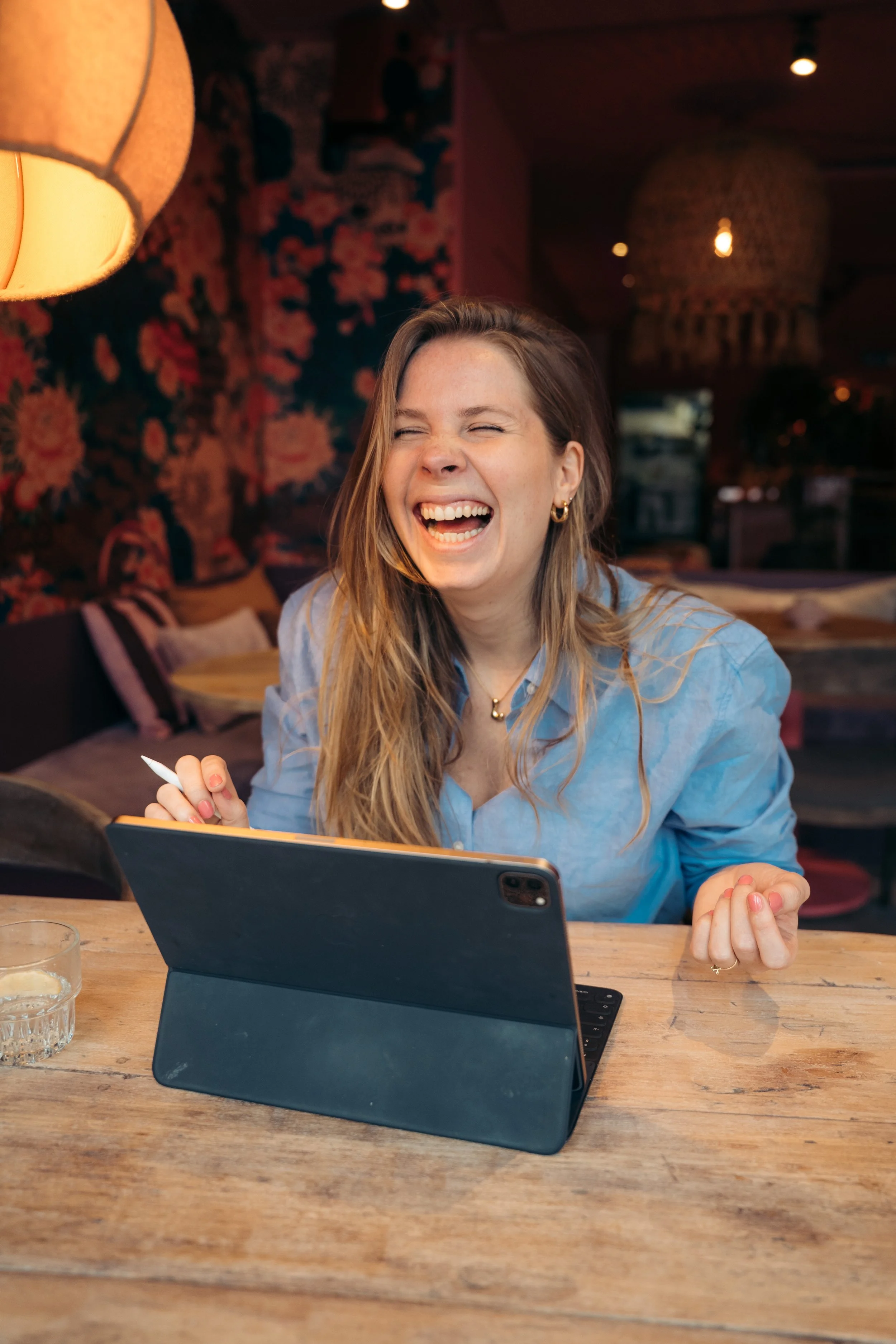 Vrolijke jonge vrouw lachen terwijl ze op een tablet in een gezellige, sfeervolle inrichting met lampen en een bloemenwand zit.