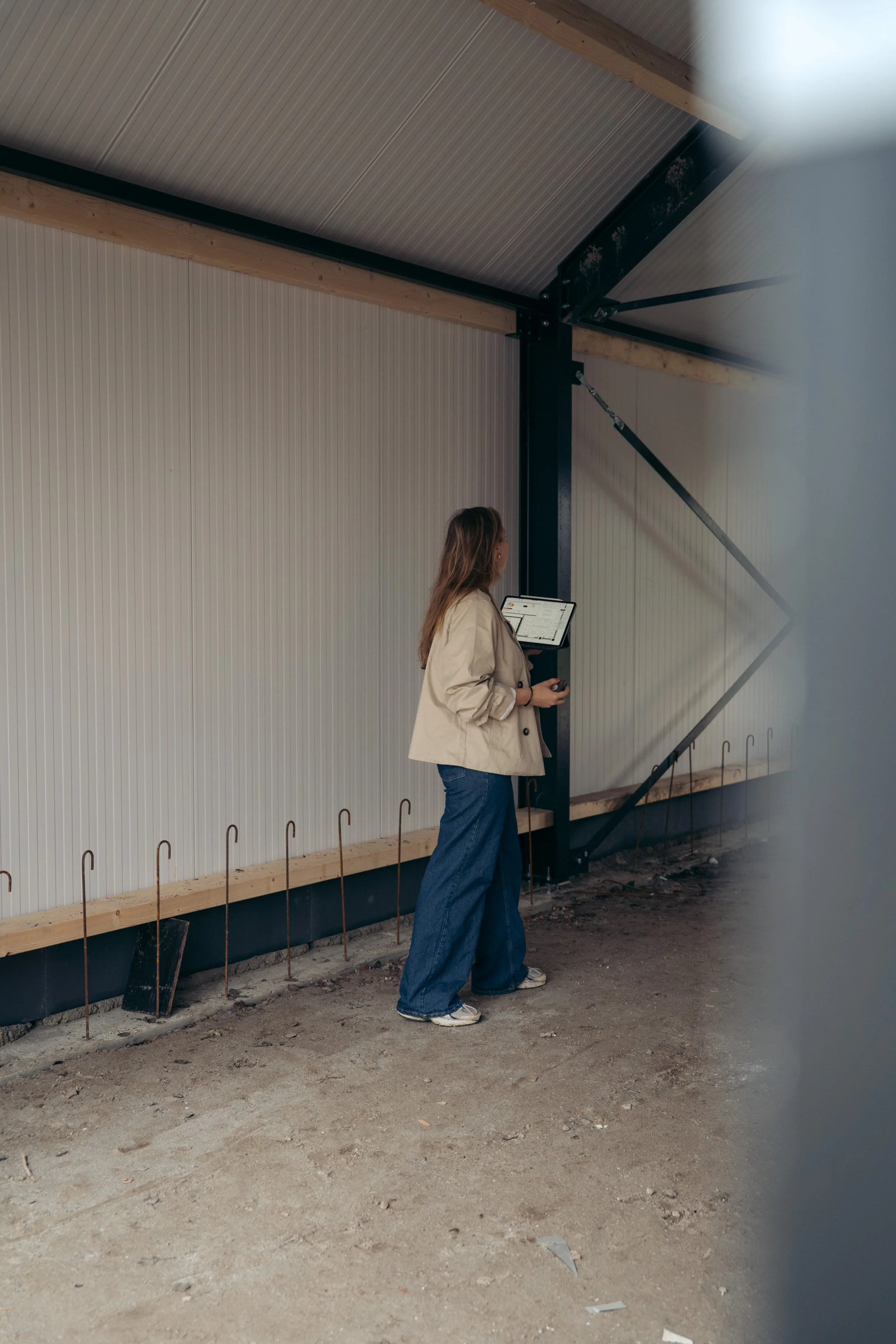 Vrouw in een beige jas en blauwe broek houdt een tablet vast terwijl ze kijkt naar een constructie in een gebouw met een metalen muur en houten balken.