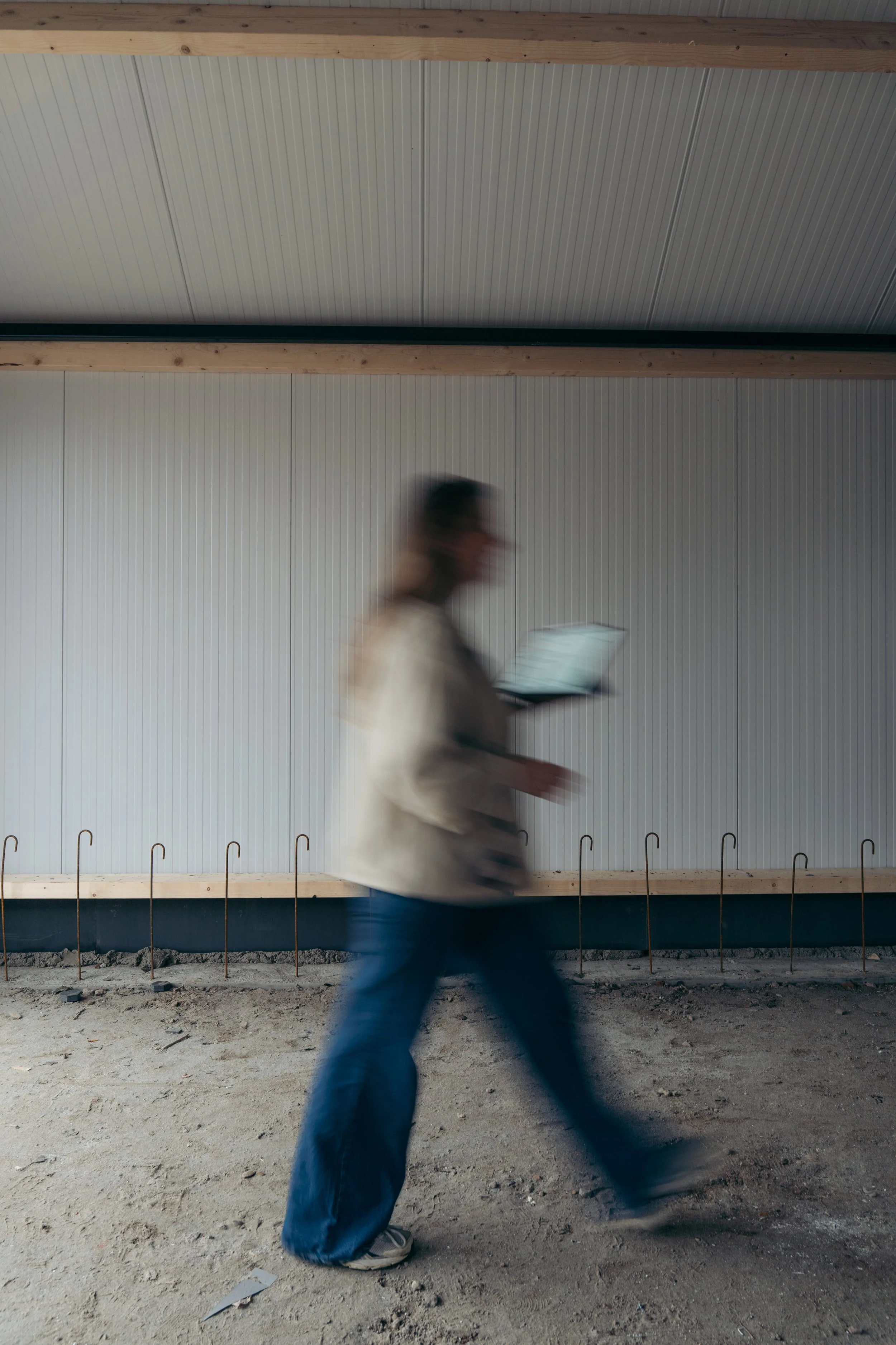 Een persoon in beweging met een witte jas en blauwe jeans loopt van rechts naar links voor een achtergrond van een bouwmuur met verticale metalen platen en een houten rand.