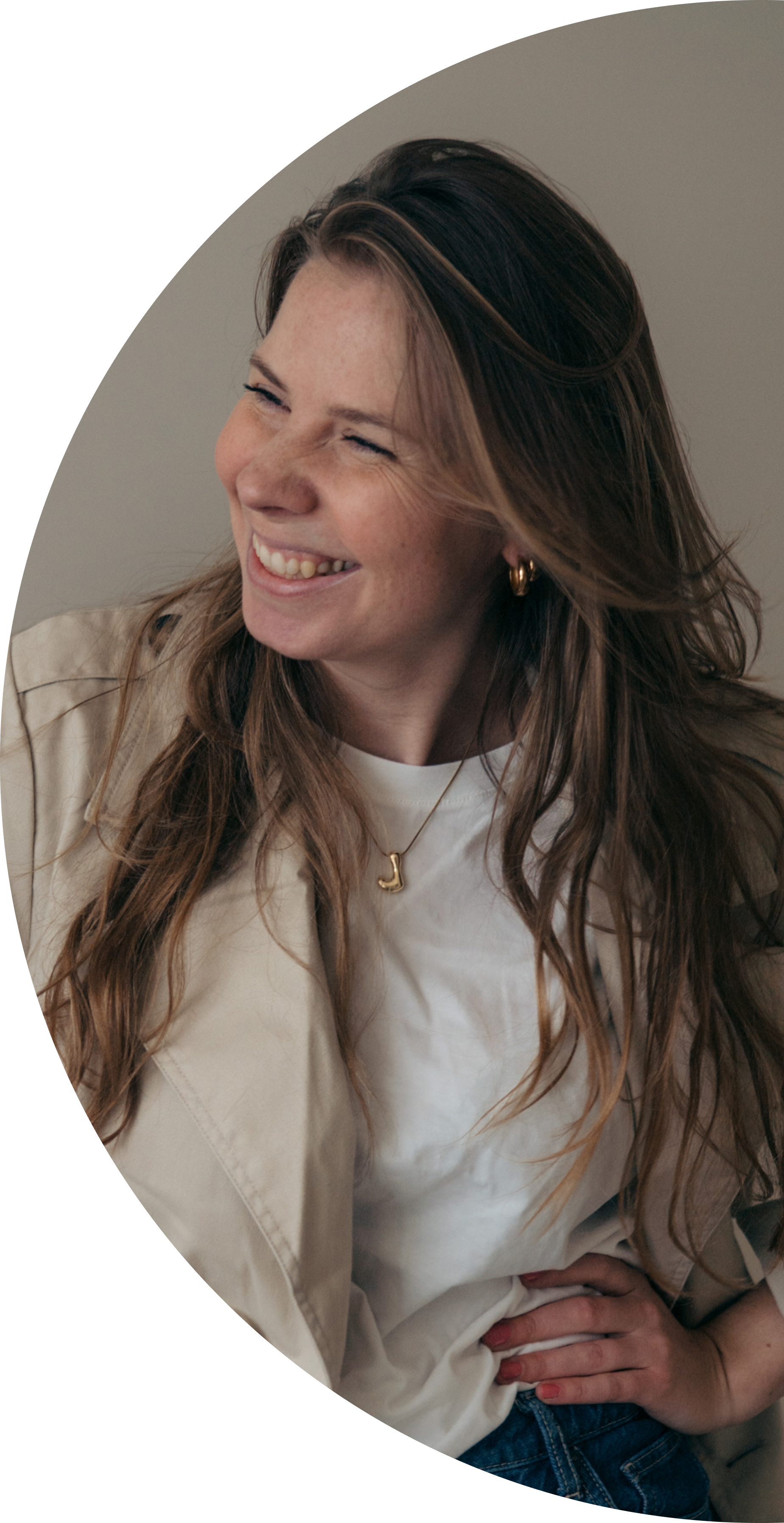 Een lachende vrouw met lang bruin haar, oorbellen en een ketting met een letter 'J', gekleed in een witte top en beige blazer, zittend en lachend.