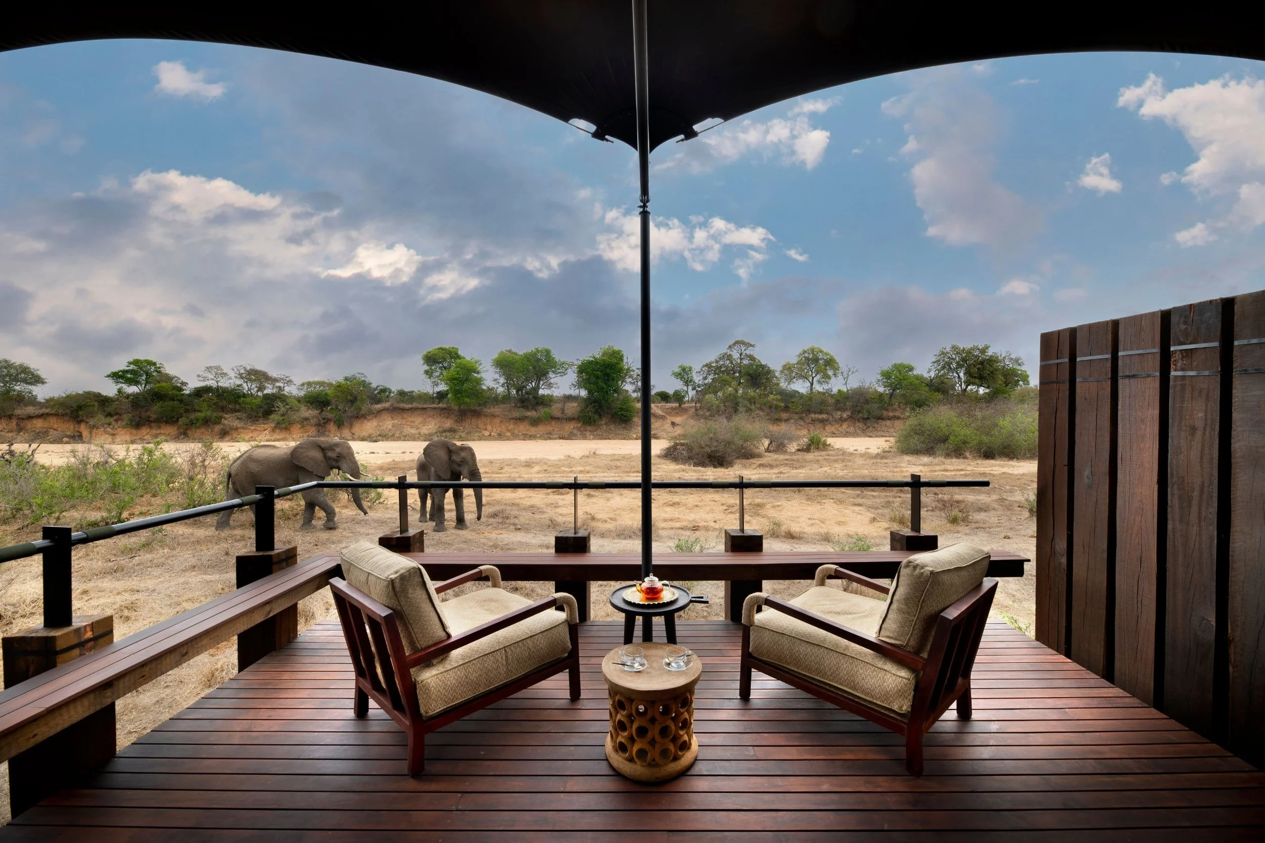 A wooden balcony with cushioned chairs and a small round table, overlooking an African savanna with three elephants walking in the distance and a bright blue sky with scattered clouds.