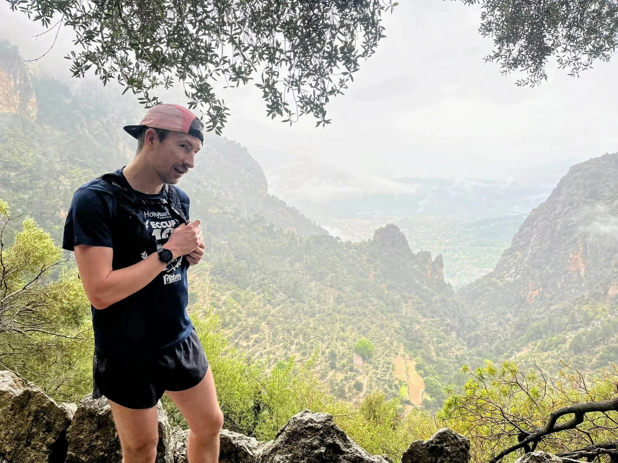 A man standing on rocky terrain in a mountainous area with lush green trees and foggy sky, wearing a black shirt, black shorts, a pink cap, and a watch, smiling while looking down, with a scenic mountain view in the background.