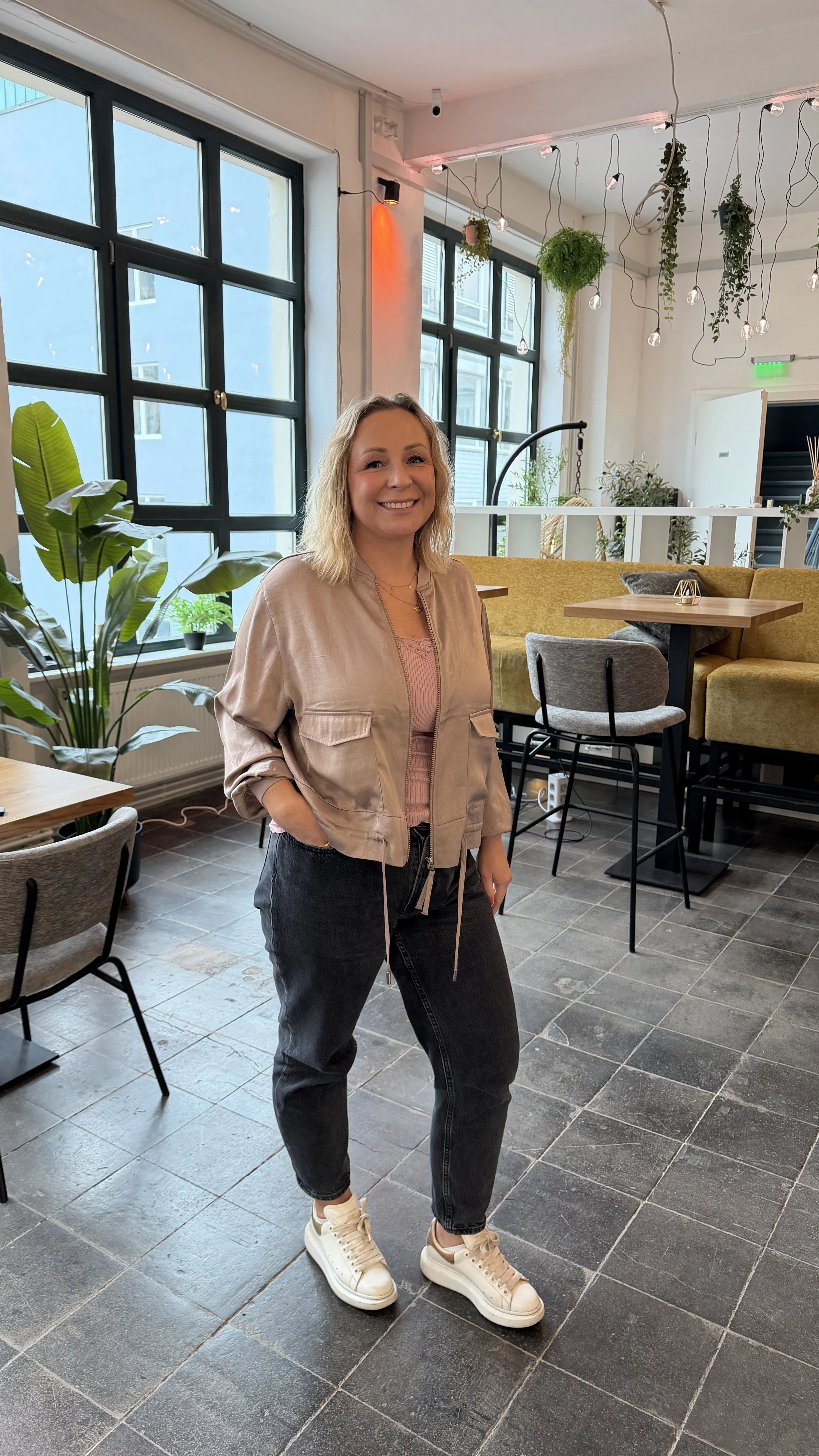 Eine lachende Frau in einem Café mit großen Fenstern, Pflanzen und moderner Inneneinrichtung.