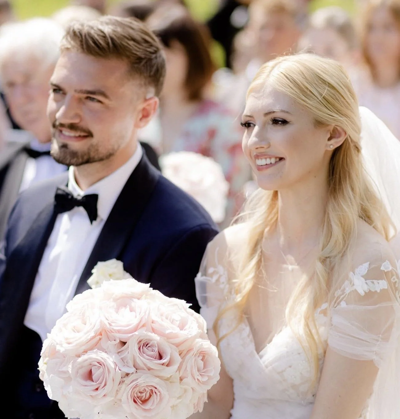 Braut und Bräutigam bei einer Hochzeitsfeier, die Braut hält einen Blumenstrauß aus hellrosa Rosen, beide sind in festlicher Kleidung, draußen bei sonnigem Wetter.