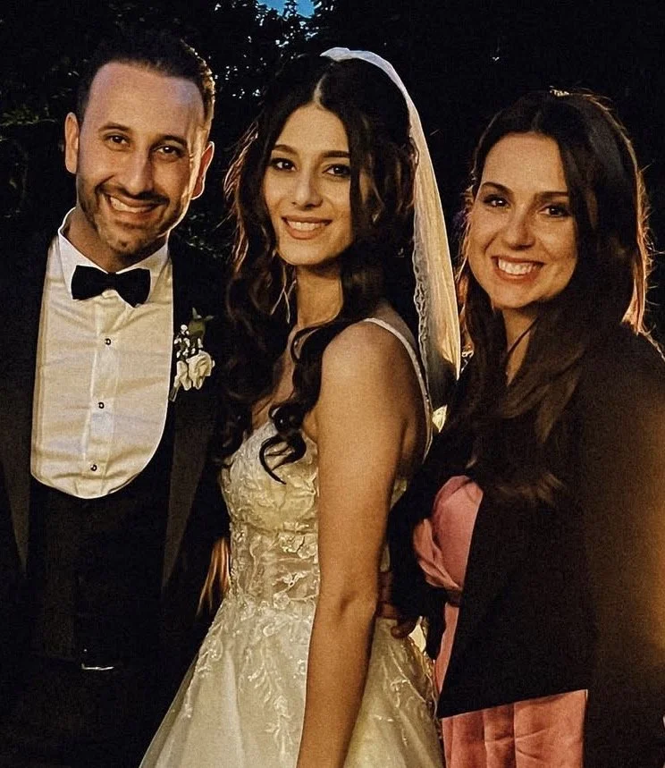 Drei Menschen auf einer Hochzeit, eine Braut in einem weißen Brautkleid mit Schleier, ein Bräutigam in einem schwarzen Anzug mit Fliege, und eine Frau im schwarzen Kleid mit rosa Akzent, alle lächelnd in der Abenddämmerung im Freien.