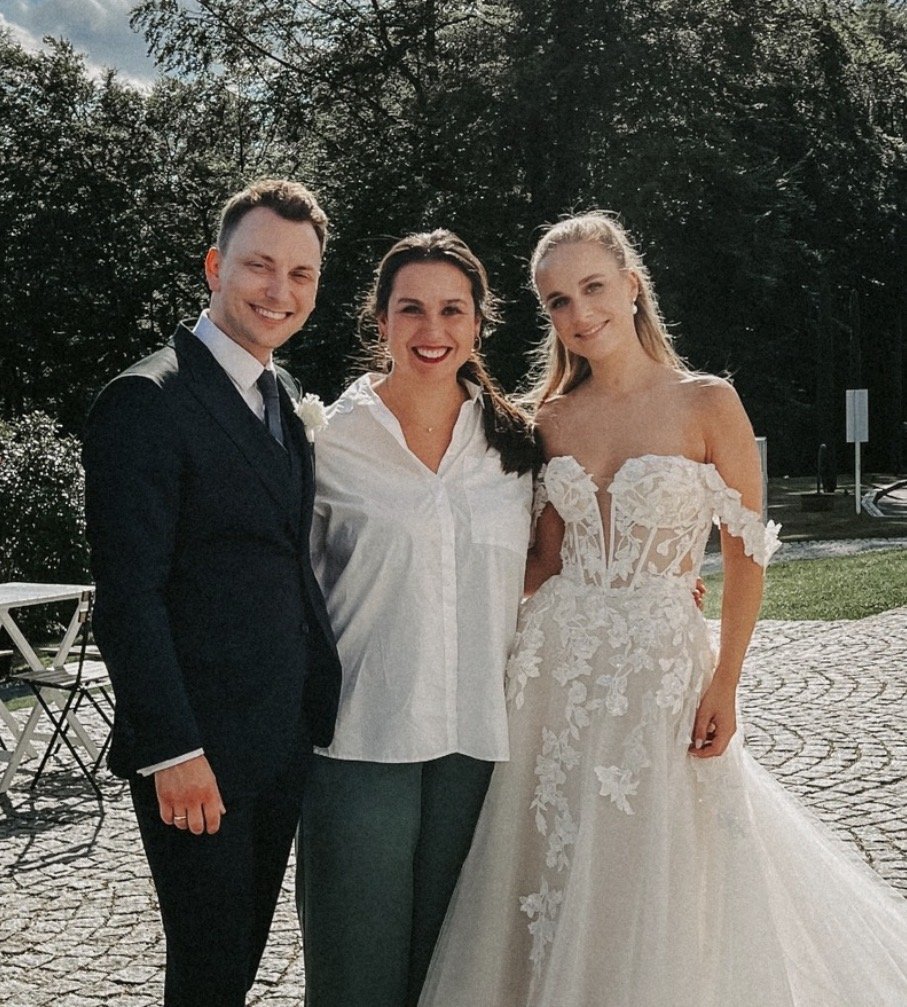 Drei Personen bei einer Hochzeit im Freien, zwei Frauen und ein Mann, der Mann trägt einen dunklen Anzug, die Frauen tragen Hochzeitskleider, die eine mit einem weißen Bodenkleid und die andere mit einem weißen Kleid mit Blumenmuster, alle lächeln.