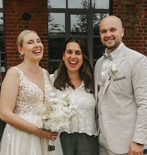 Drei Menschen auf einer Hochzeit, ein Brautpaar und eine Frau zwischen ihnen, alle lachen und posieren vor einem Fenster mit Backsteinmauer.