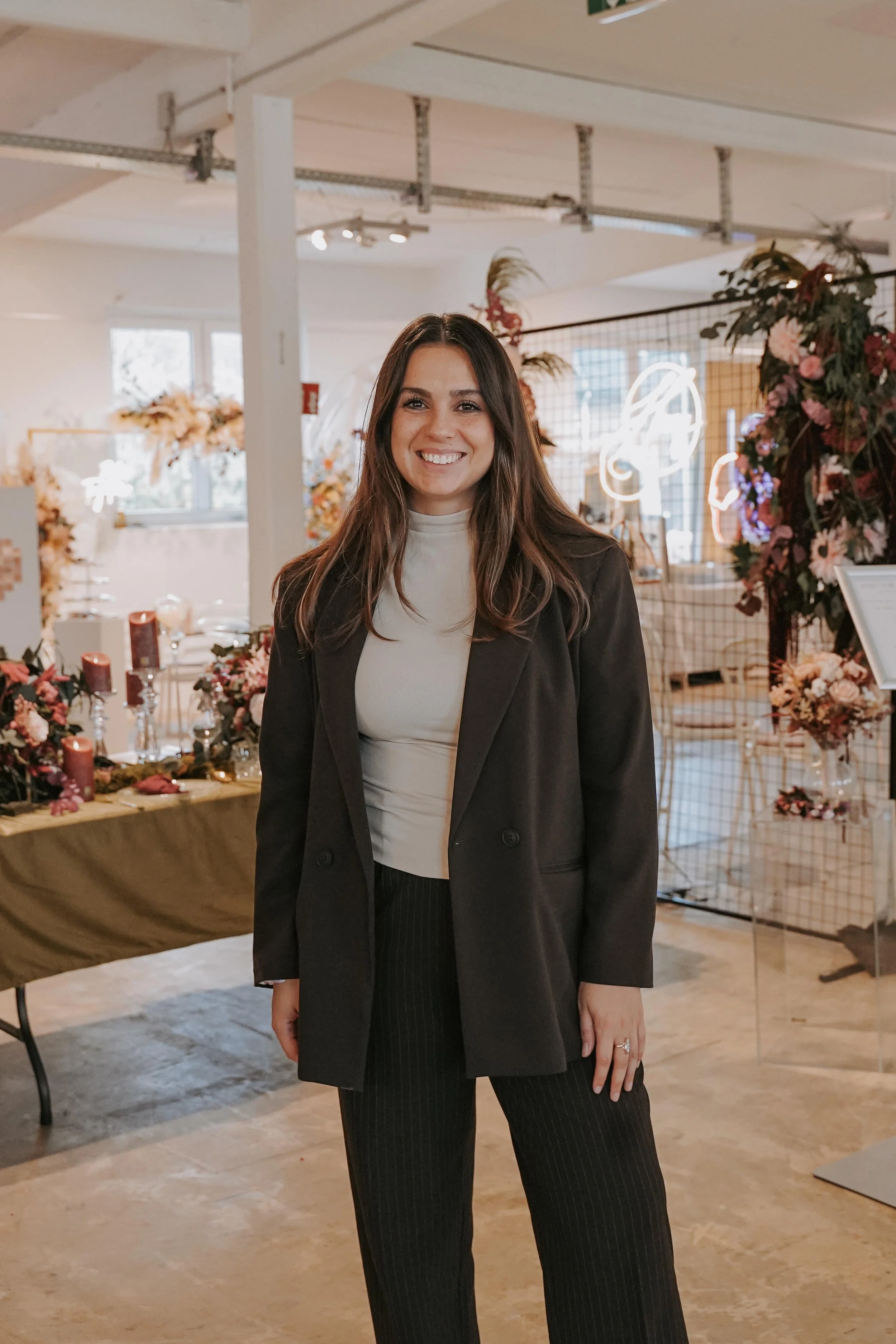 Eine lächelnde Frau trägt einen schwarzen Blazer und eine weiße Bluse in einem dekorierten Raum mit Blumen und Neonlichtern.