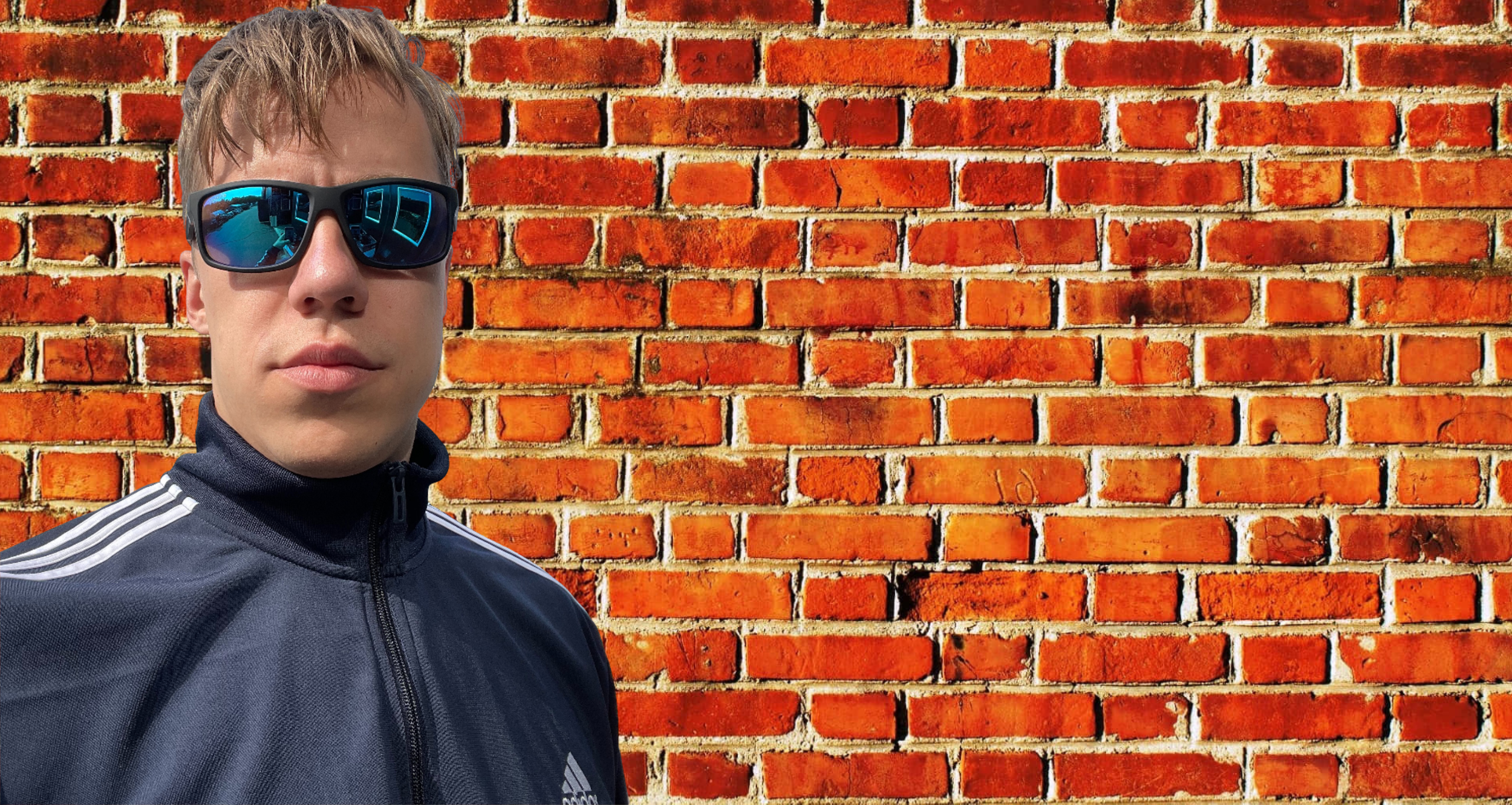 A young man wearing sunglasses with blue mirrored lenses and a dark sports jacket with white stripes on the shoulders, standing in front of a red brick wall.