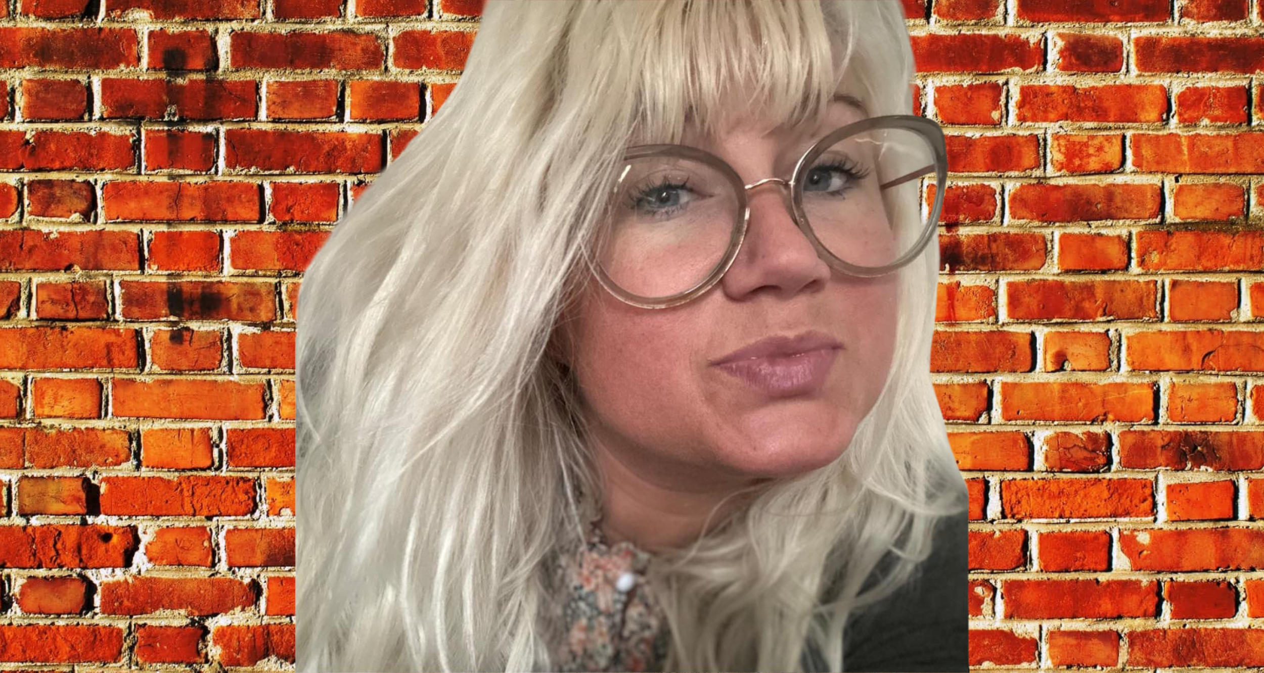A woman with long blonde hair, wearing large round glasses, posing in front of a red brick wall.