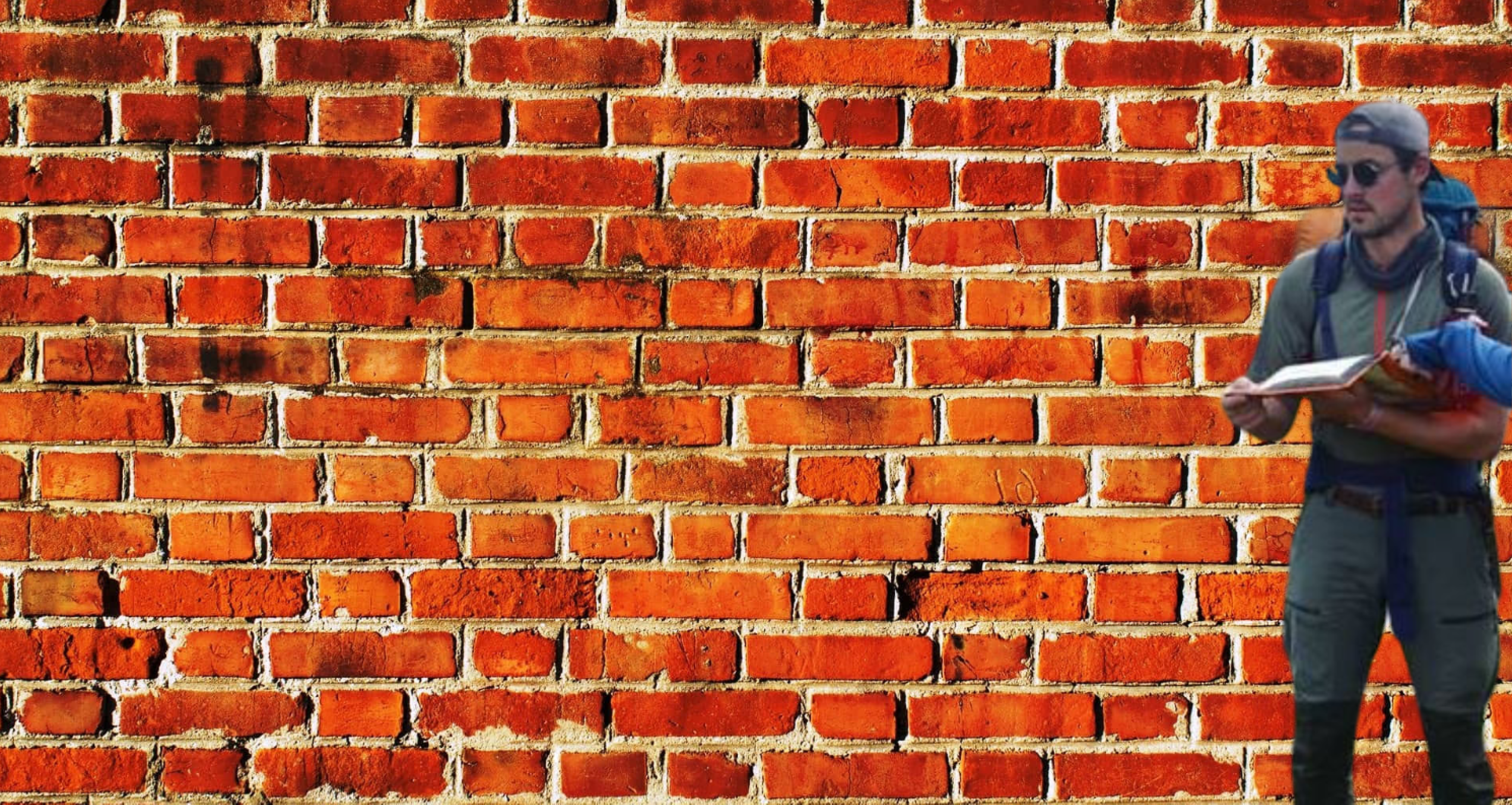 A person with sunglasses, a gray cap, and a backpack, holding a map or notebook, standing next to a red brick wall.