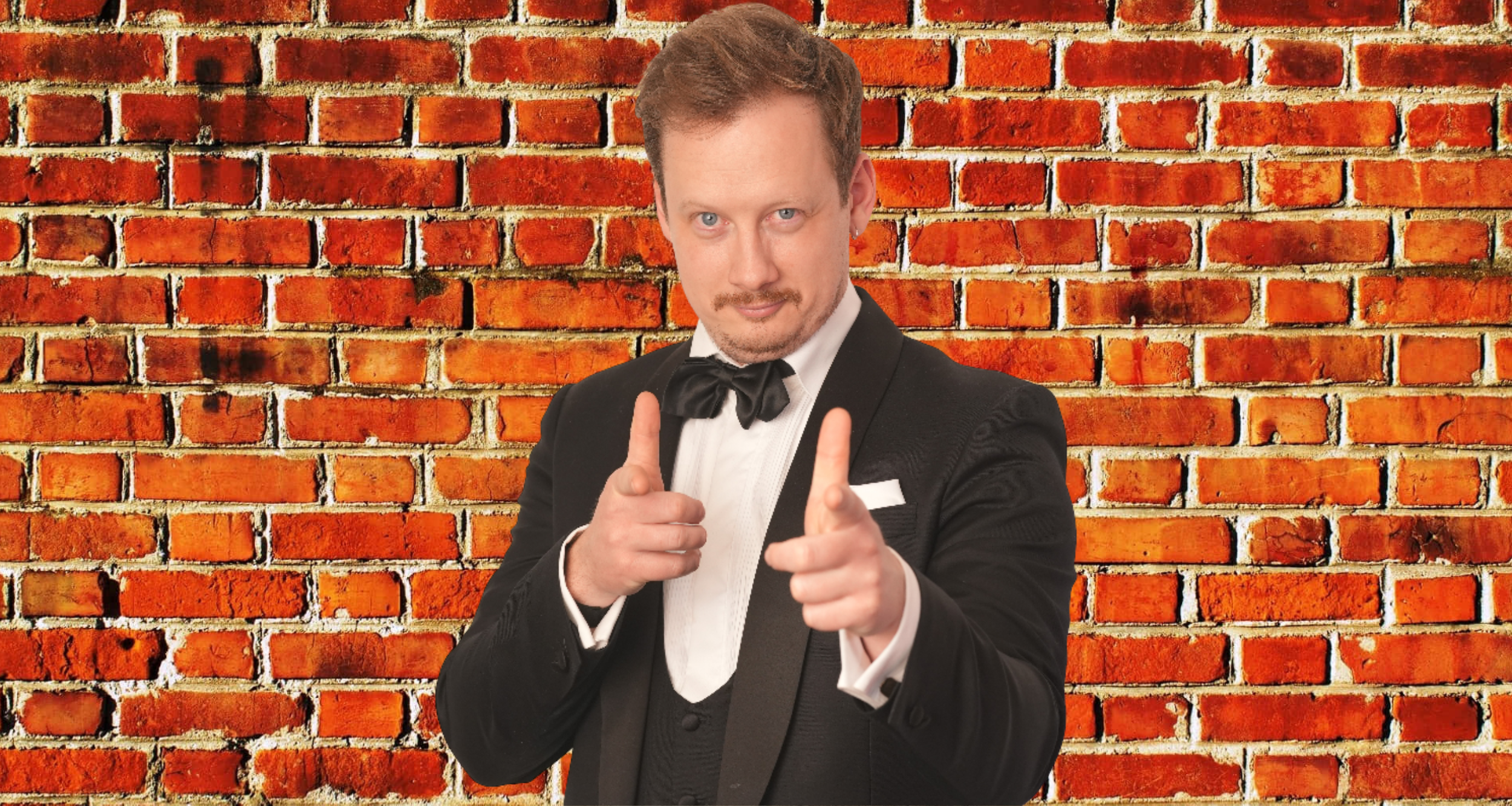 A man in a tuxedo with a black bow tie and white shirt pointing directly at the camera against a backdrop of a red brick wall.