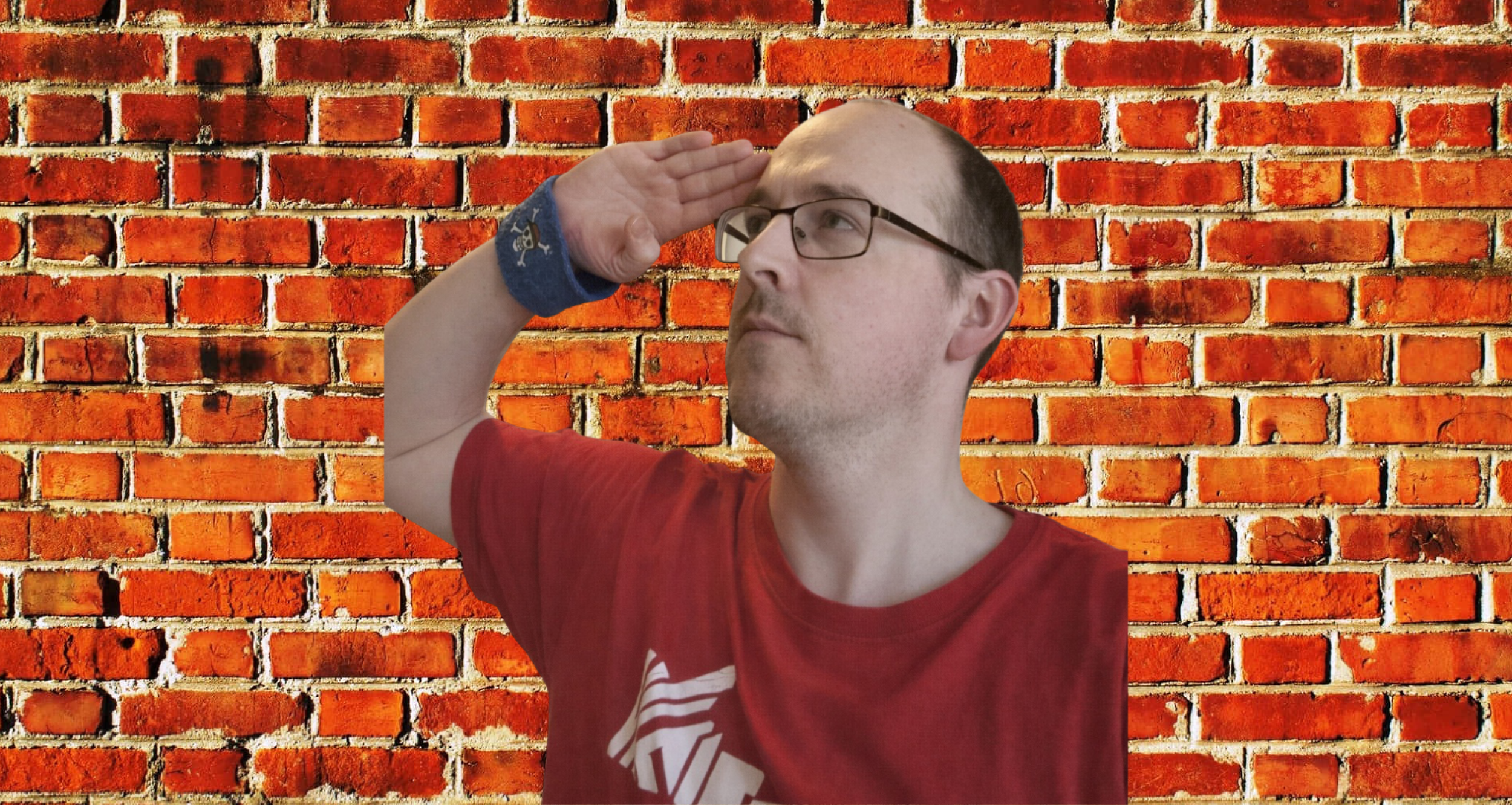 A man with glasses wearing a red shirt and a blue bracelet salutes against a brick wall.