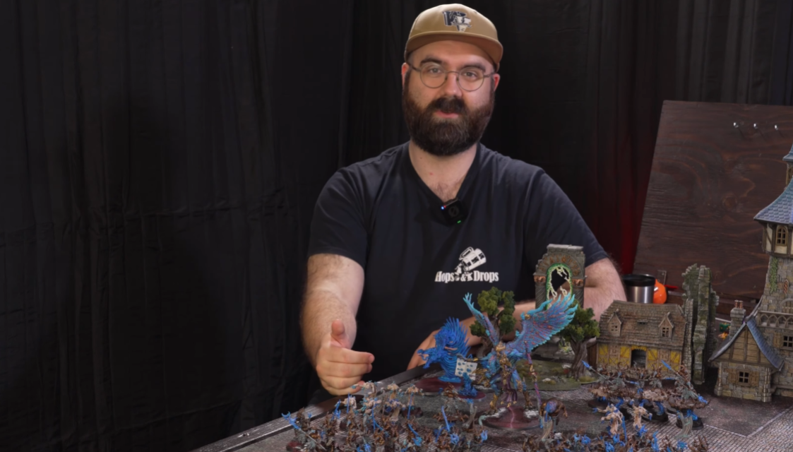 A man with glasses, a beard, and a tan cap, sitting at a table with intricate miniature models and terrain for tabletop gaming, giving a thumbs up.