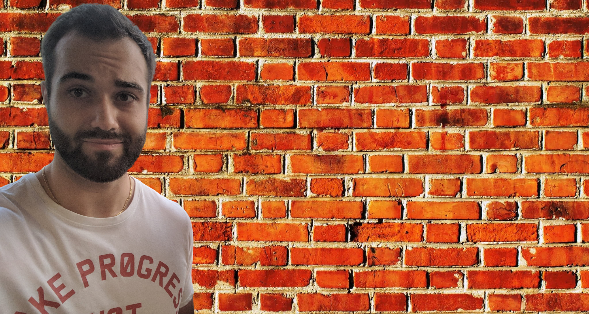 A man with dark hair and a beard taking a selfie in front of a brick wall. He is wearing a white T-shirt with red text