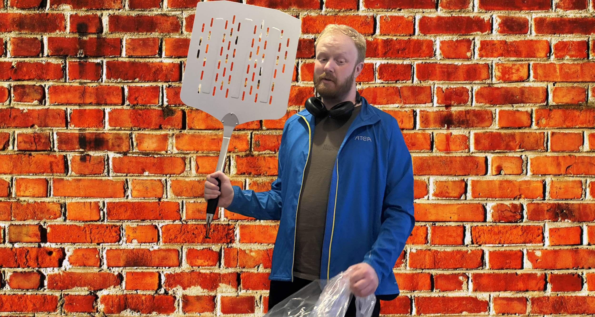 A man with red hair and a beard wearing a blue jacket, standing in front of a red brick wall, holding a large slotted spatula in one hand and a plastic bag in the other, with black headphones around his neck.