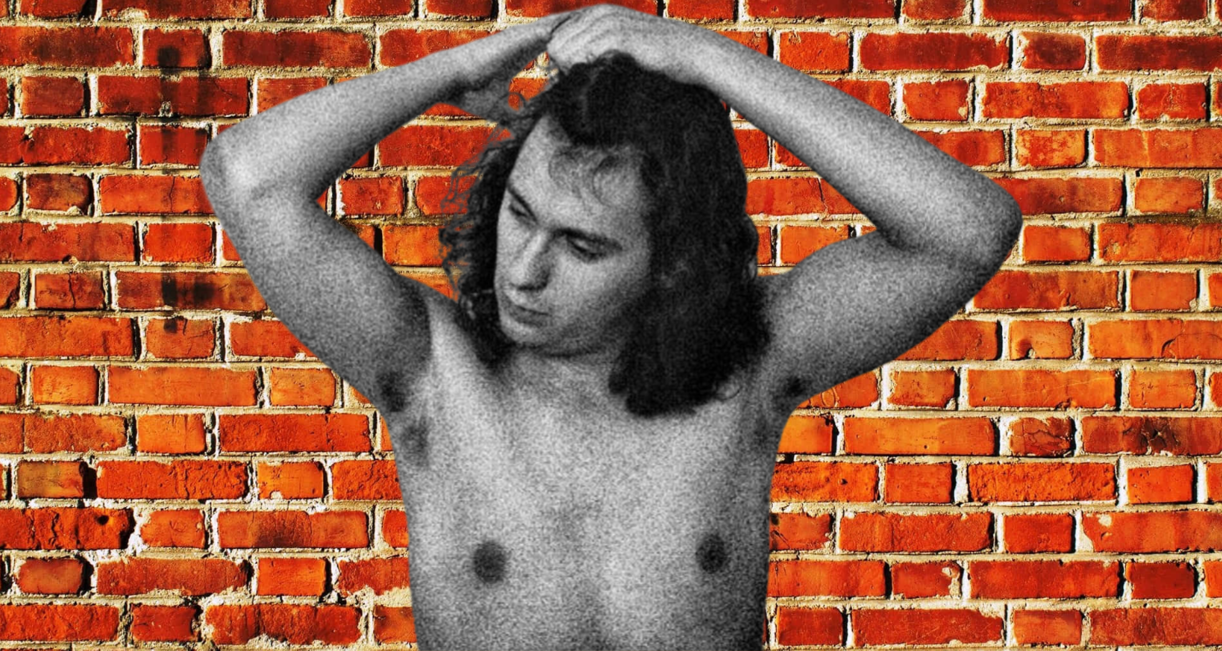 A black and white image of a topless man with long curly hair, standing in front of a brick wall, with his arms raised above his head and hands clasped behind his head.