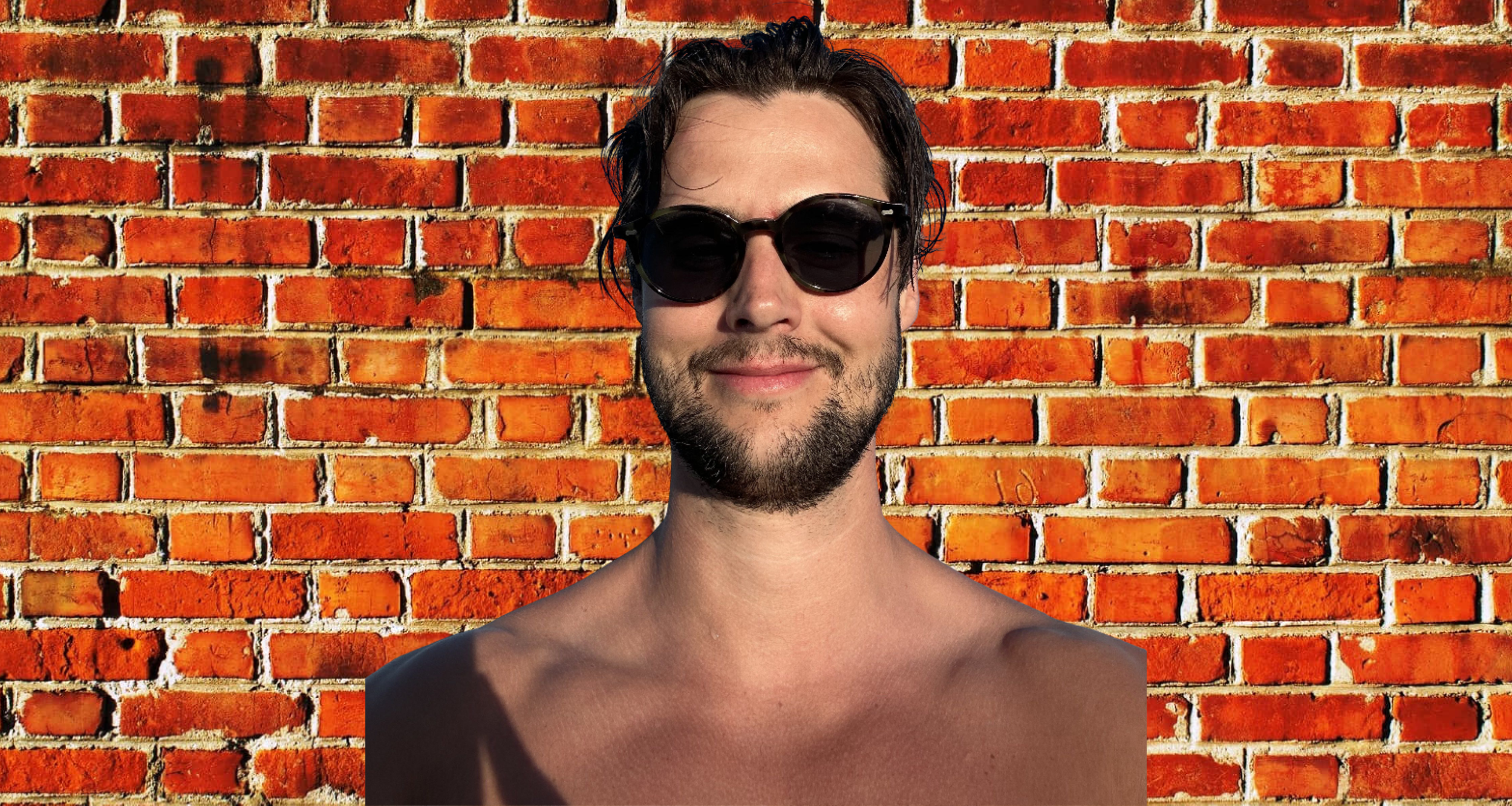 A shirtless man with dark haircut, beard, and sunglasses smiling in front of a red brick wall.