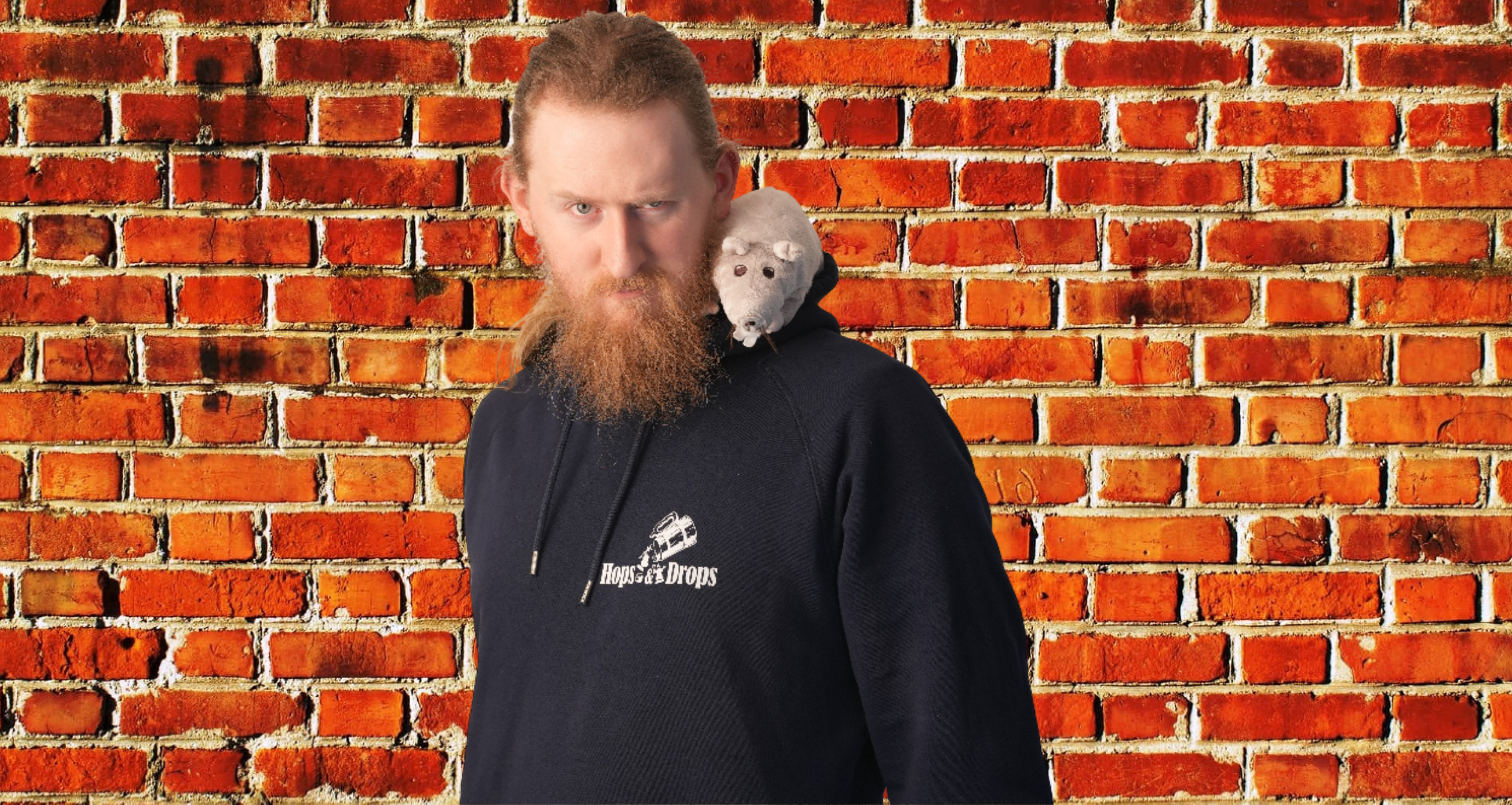 A man with red hair and beard wearing a black hoodie with 'Hops & Drops' logo, standing in front of a brick wall. A stuffed rat toy rests on his shoulder.