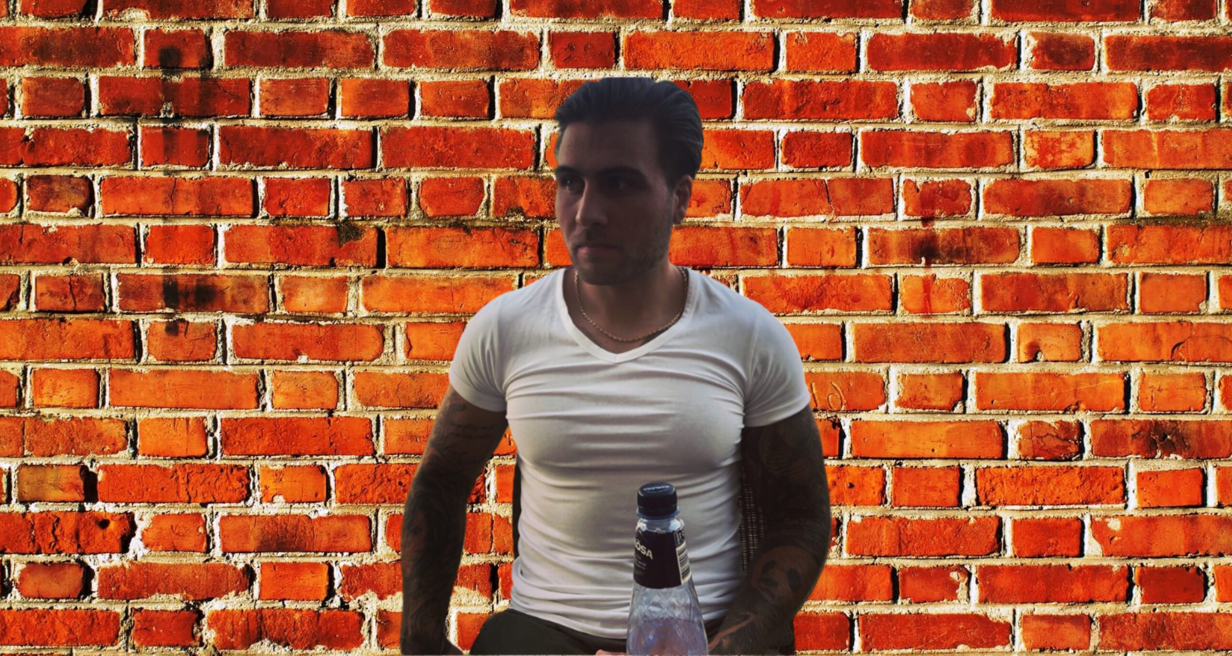 A man with tattoos on his arms wearing a white t-shirt and a gold chain, sitting in front of a red brick wall background, with a bottle of water in front of him.