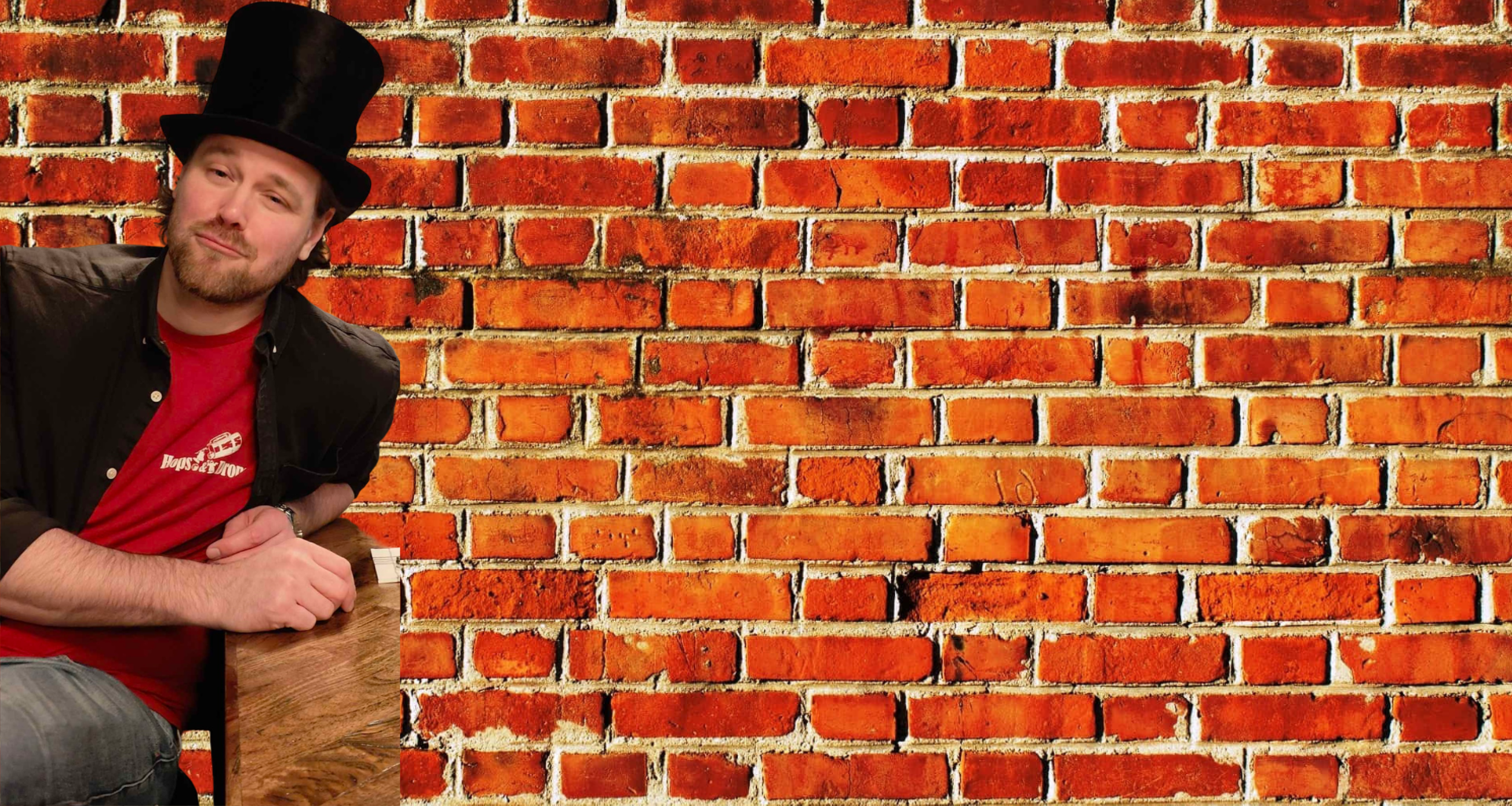 A man wearing a black top hat, black jacket, and red T-shirt sitting at a wooden table in front of a brick wall.