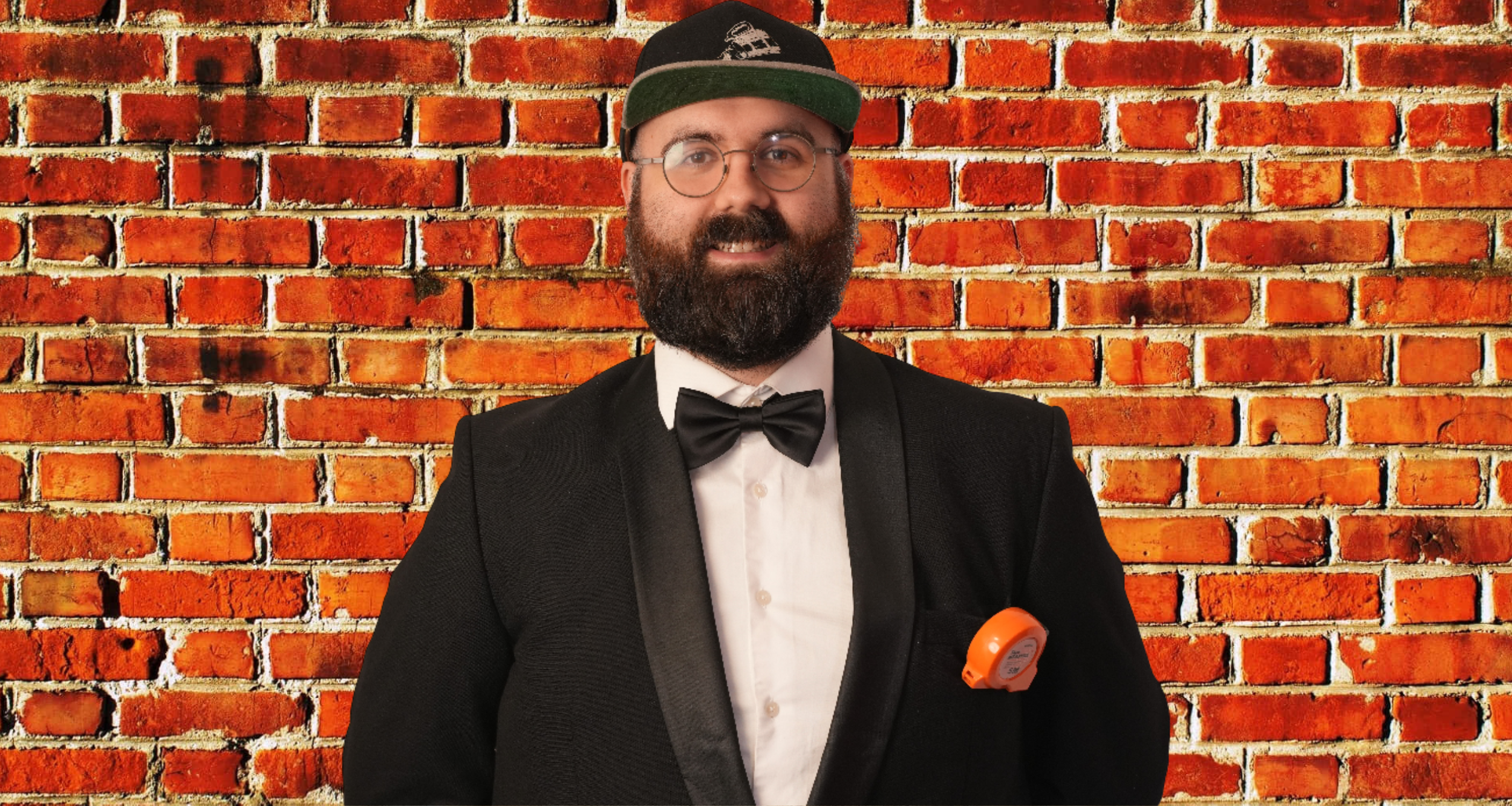 A man with glasses, a beard, and a mustache wearing a black tuxedo with a white dress shirt and black bow tie, standing in front of a brick wall. He is also wearing a black cap with a logo and has an orange device clipped to his tuxedo pocket.