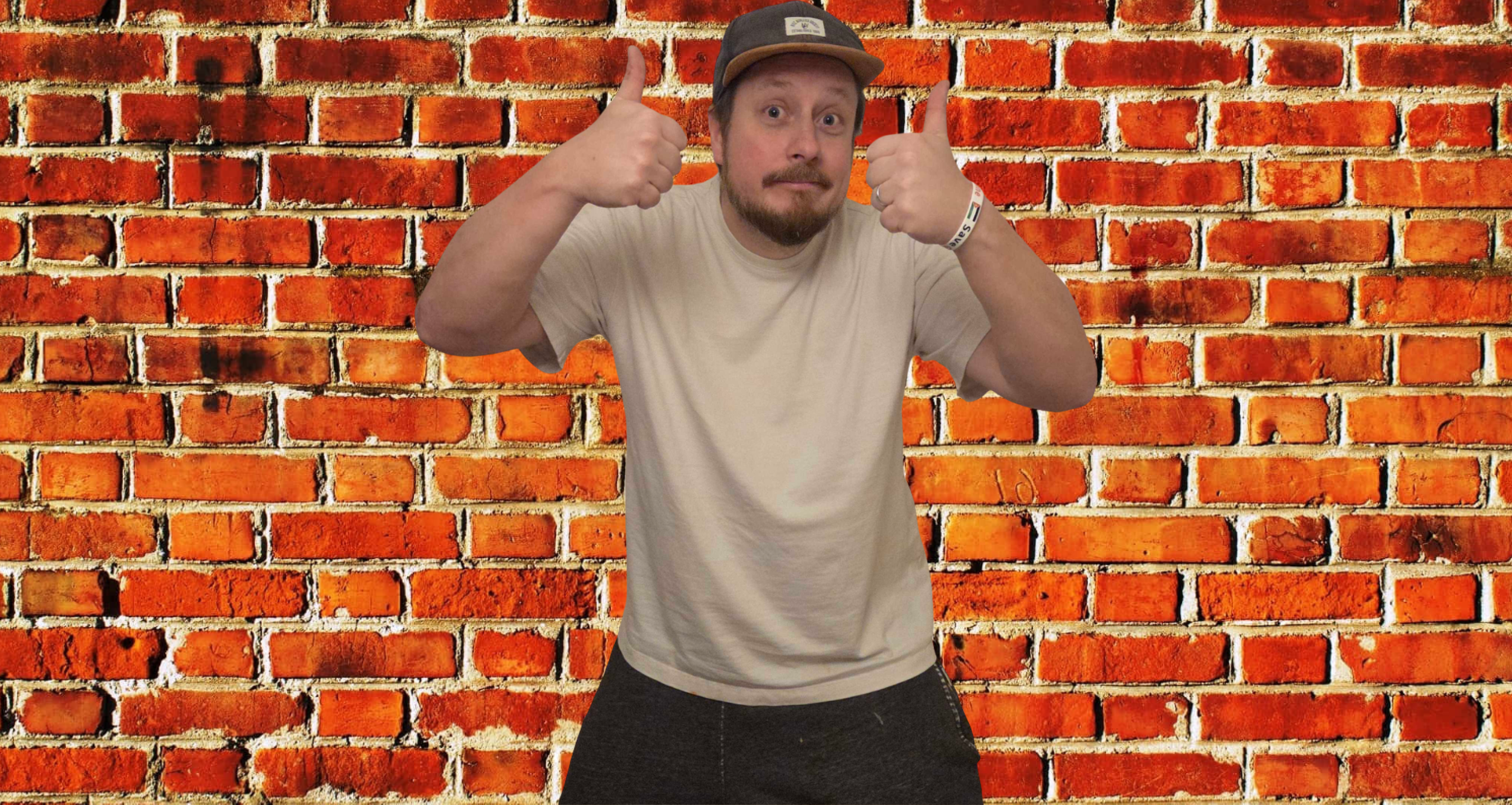 Man with a beard and mustache wearing a beige t-shirt, dark pants, and a cap, standing in front of a red brick wall. He is giving two thumbs up and looking at the camera with a slight smile.
