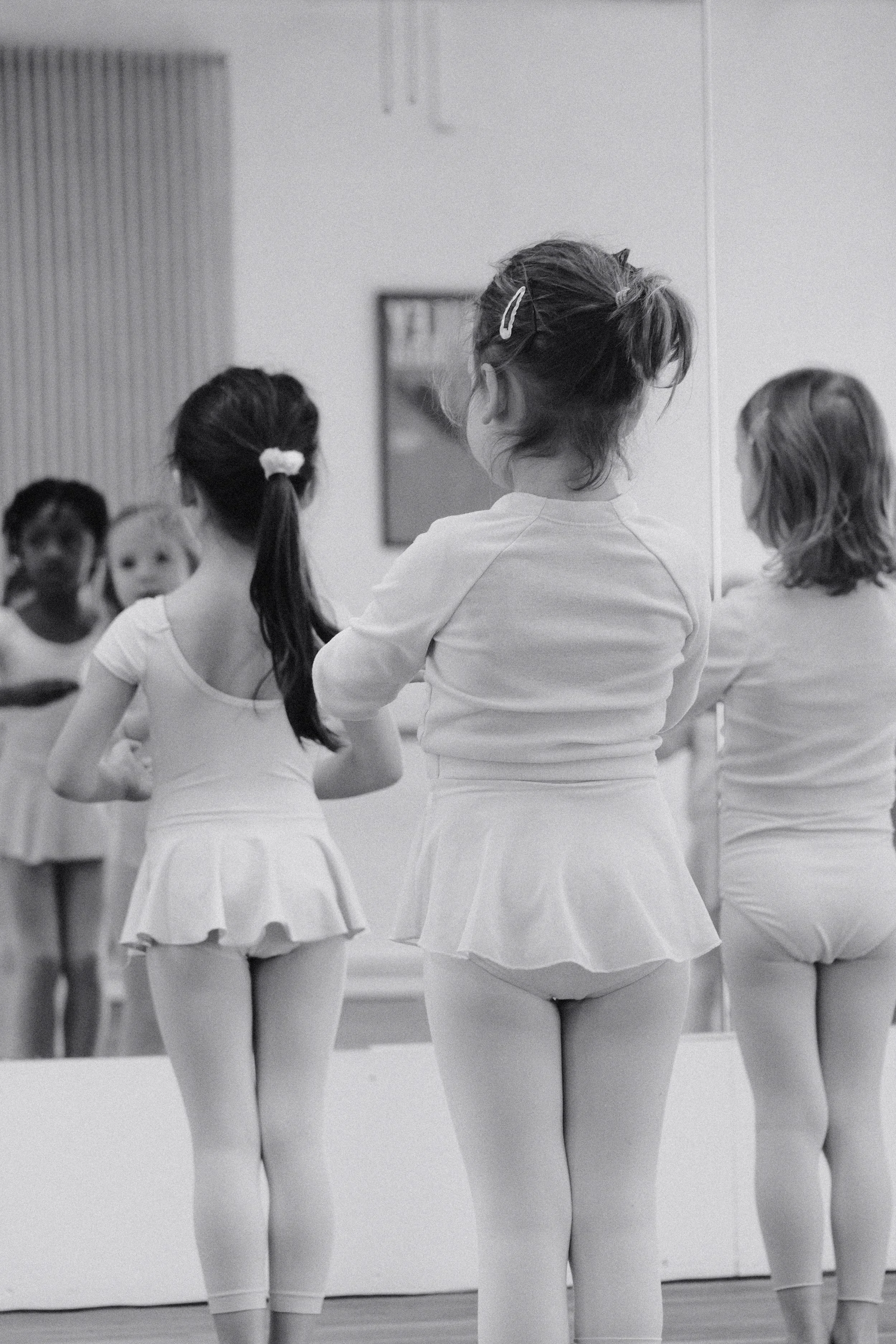 Trois jeunes filles en tenues de danse regardent leur reflet dans le miroir, l'une d'entre elles a les cheveux attachés avec une pince et porte une jupe à volants.