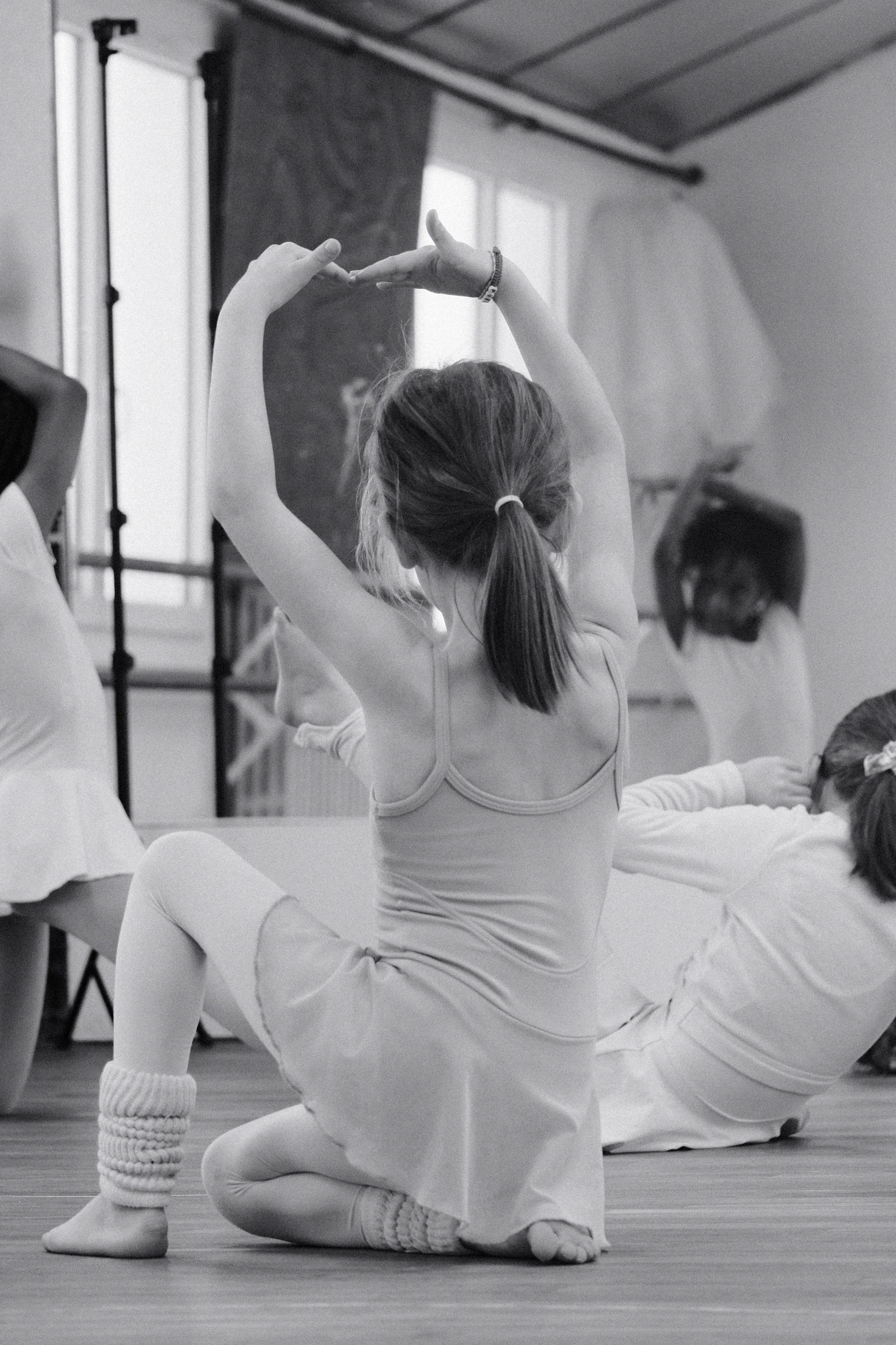 Une jeune fille en tutu pratique la danse classique dans un studio, assise au sol avec les bras levés, entourée d'autres enfants également en tenue de danse.