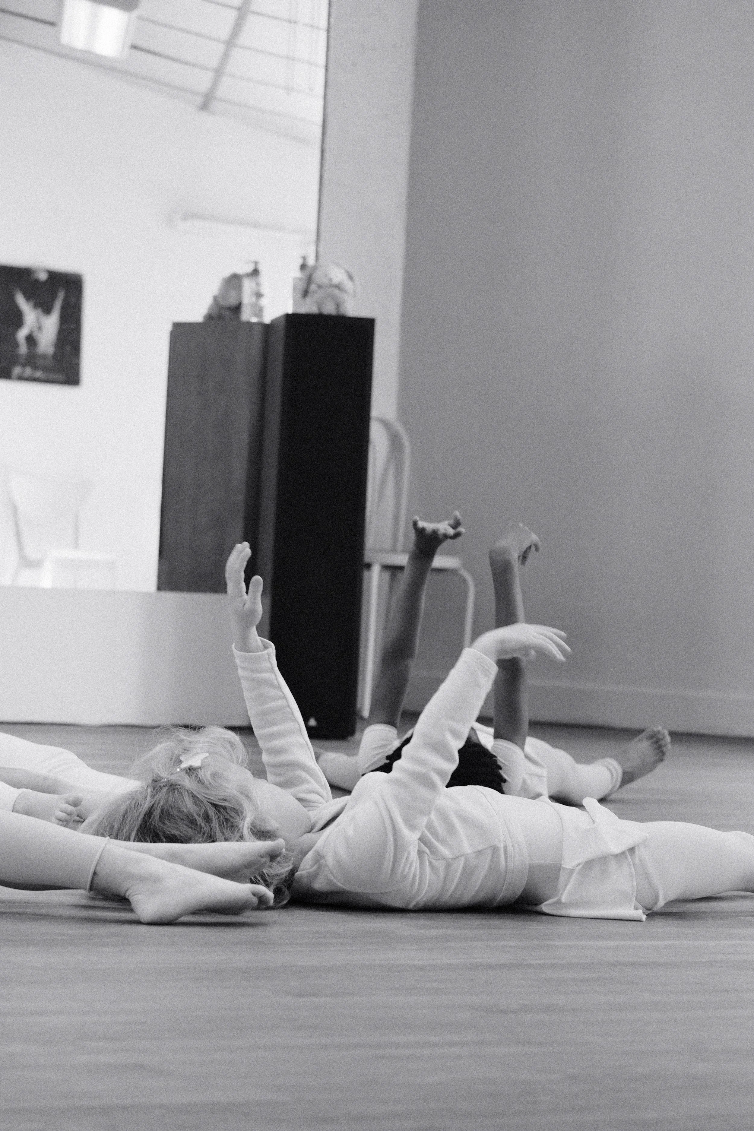 Enfants qui pratiquent la danse o au sol dans une salle, certains levant la main, en noir et blanc.