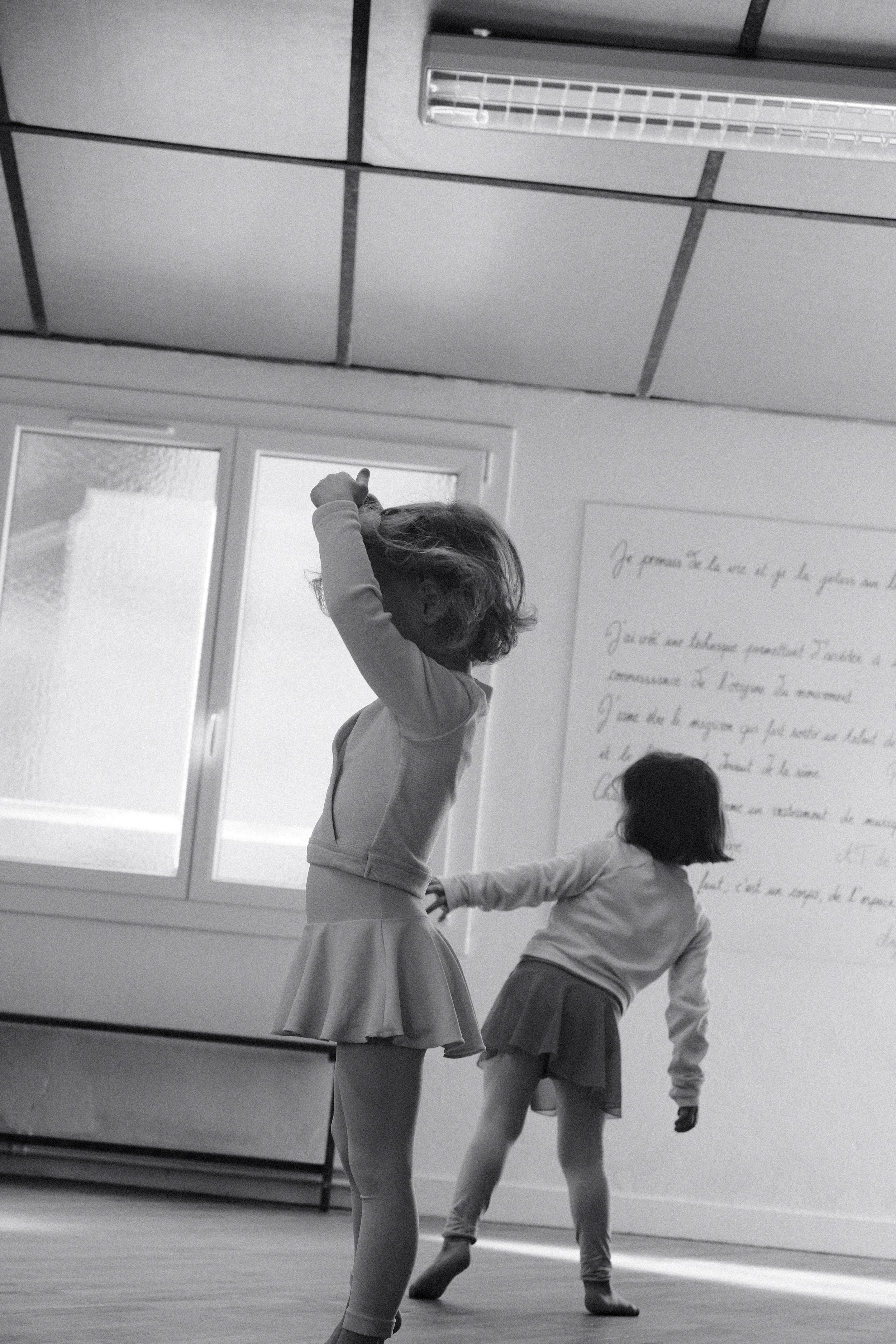 Deux jeunes filles en vêtements décontractés dans une salle, l'une avec les bras levés, l'autre en position inclinée, en train de danser , avec une fenêtre et un tableau blanc avec écritures en arrière-plan.