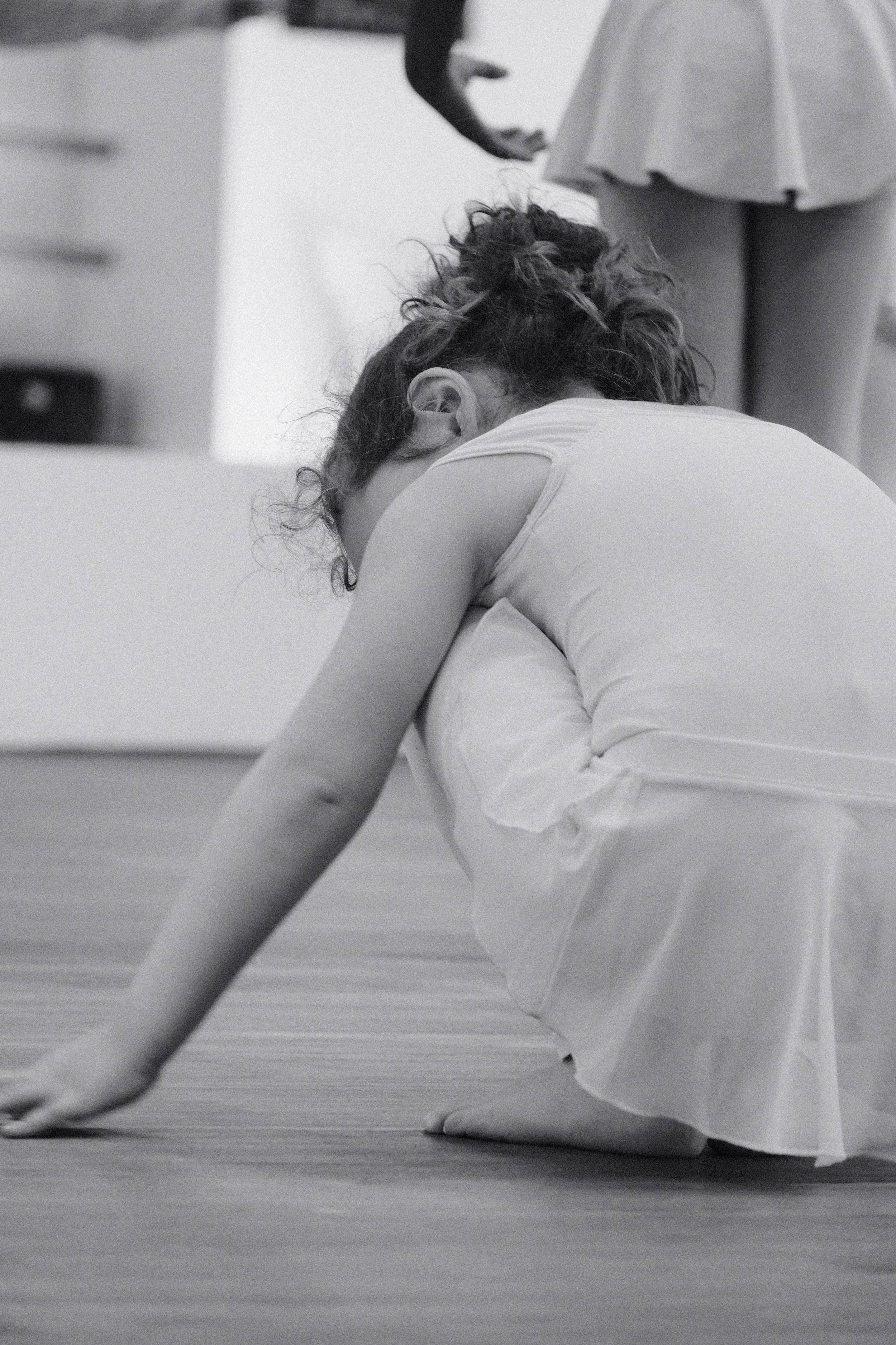 Une jeune fille en robe blanche pose sa main sur le sol du studio de danse la tête inclinée vers l'avant 