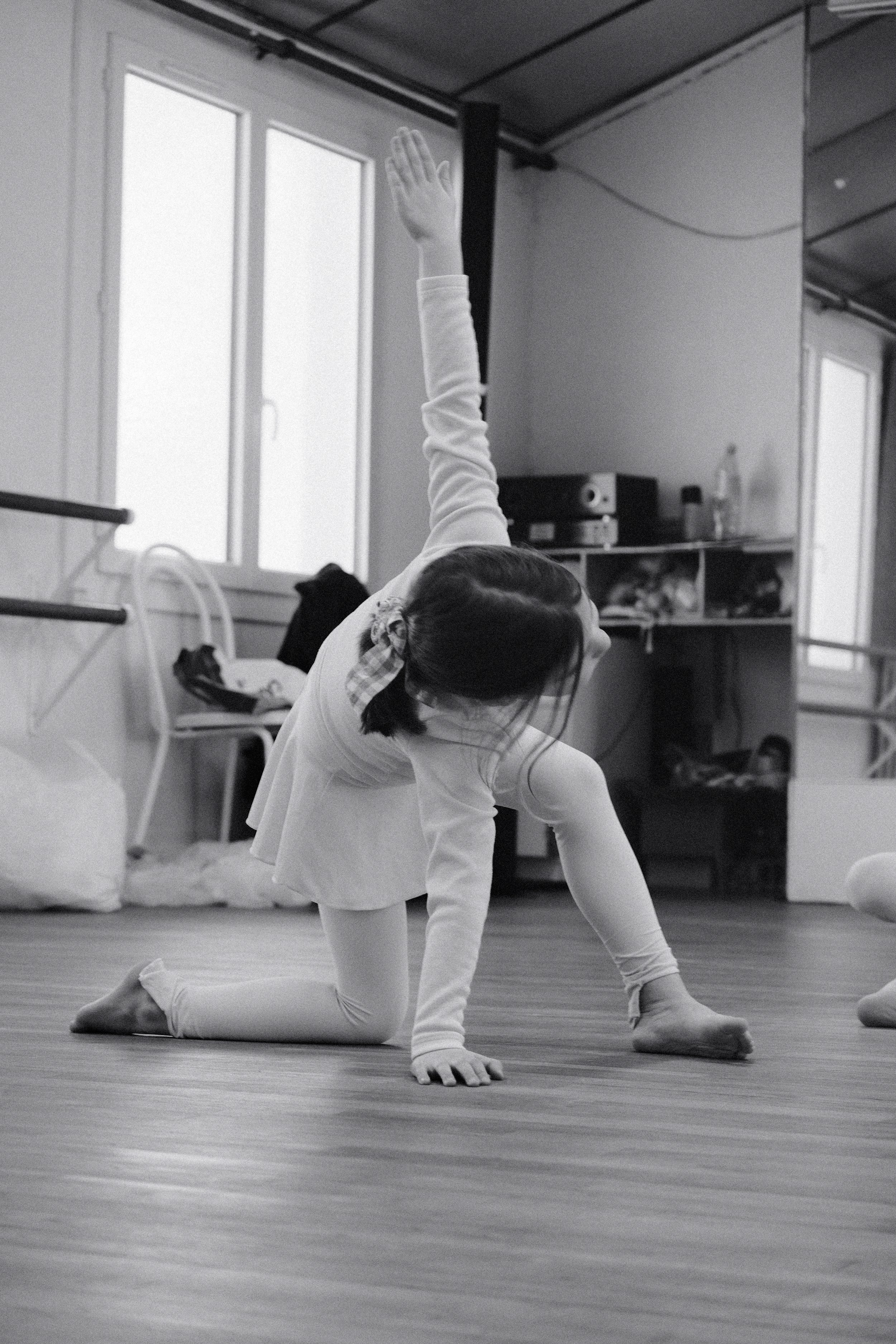 Une jeune fille pratique la gymnastique ou le yoga en position pliée avec une jambe levée, dans une salle avec des fenêtres lumineuses.