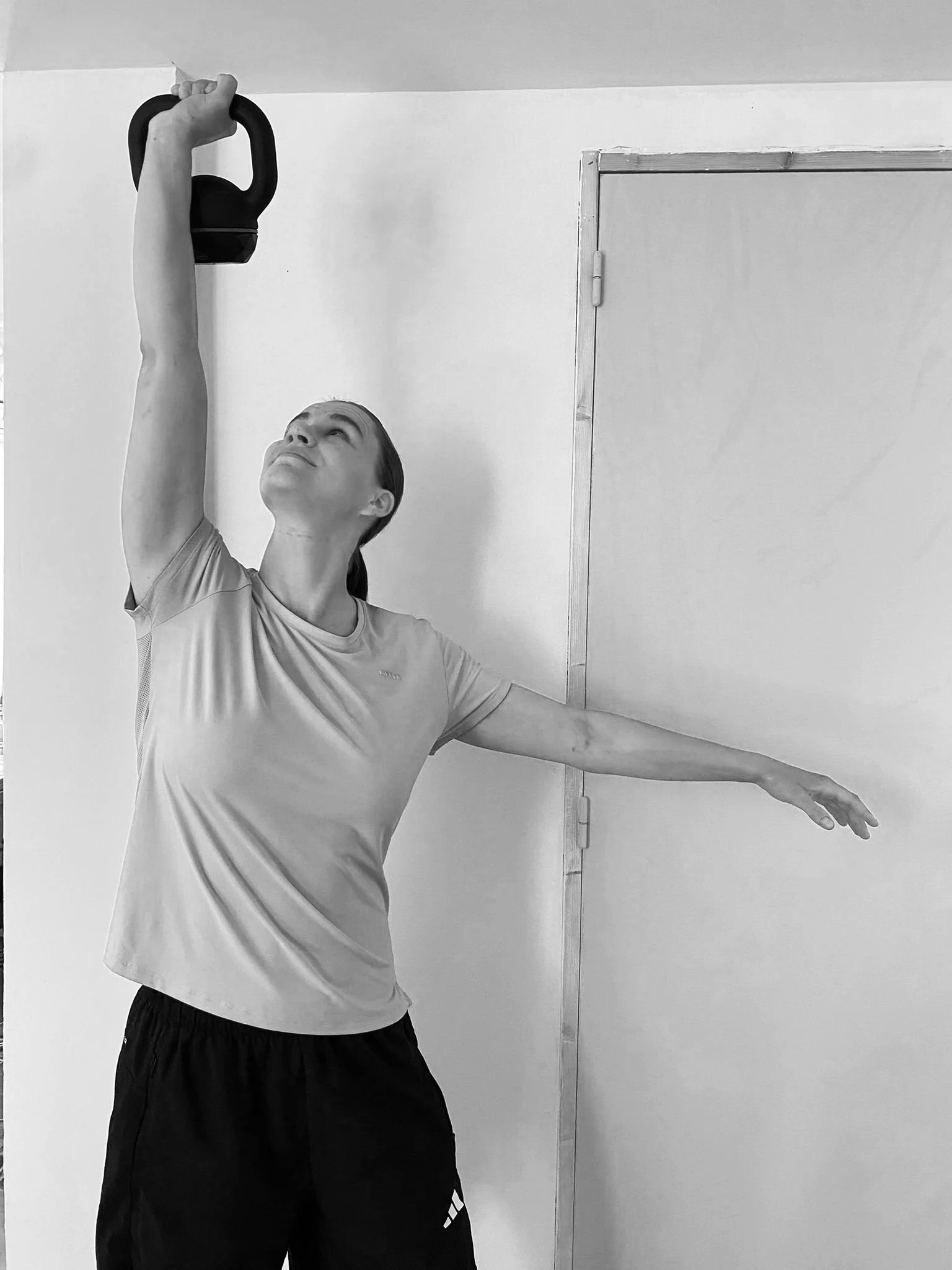 Une personne pratique le yoga ou une activité de stretching, tenant une kettlebell en l'air avec le bras gauche tendu, dans une pièce avec un mur blanc et une porte en bois.