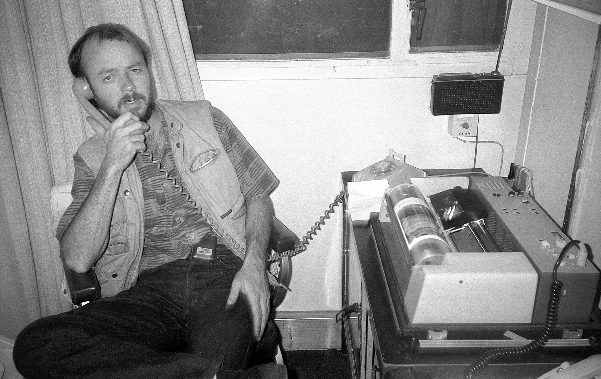 A man with a beard and mustache sitting on a chair, talking on the phone, in a room with a window and vintage electronic equipment on a desk.