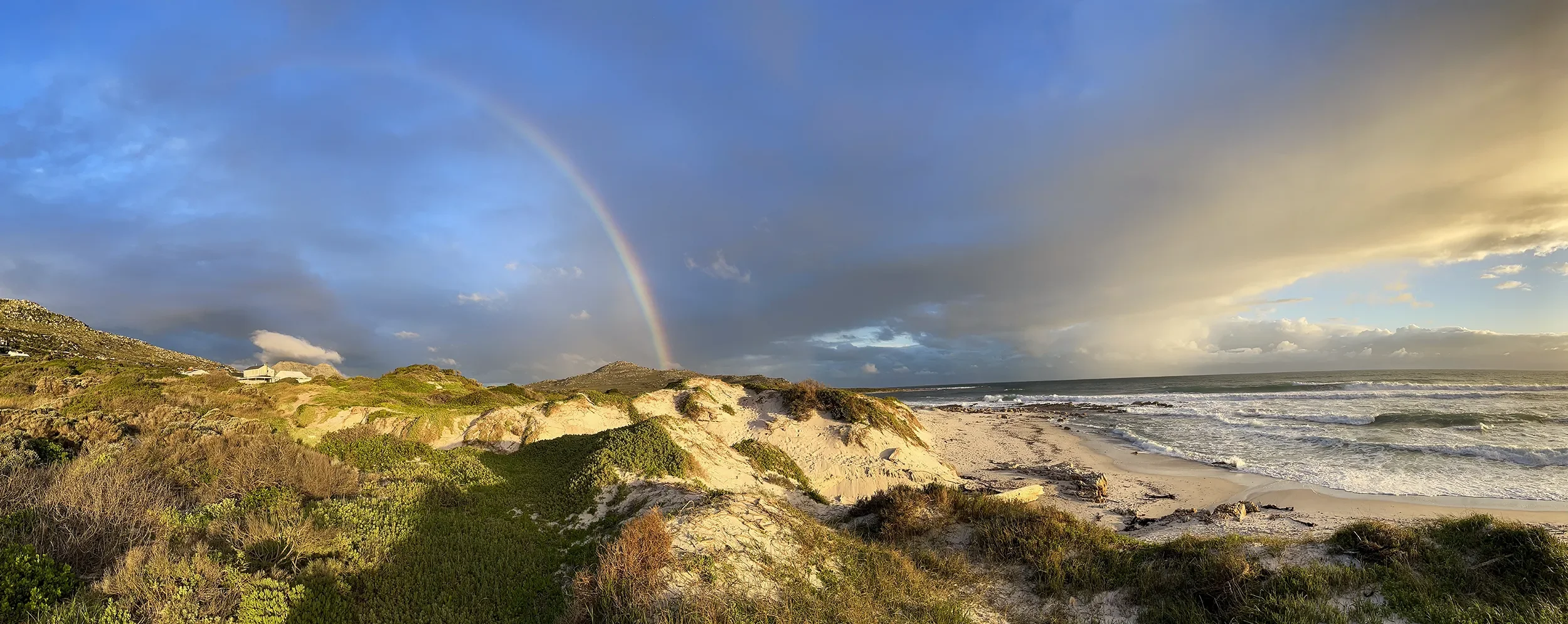 Beach scene with sand dunes covered in green vegetation, ocean waves, and a rainbow in a partly cloudy sky during sunset.