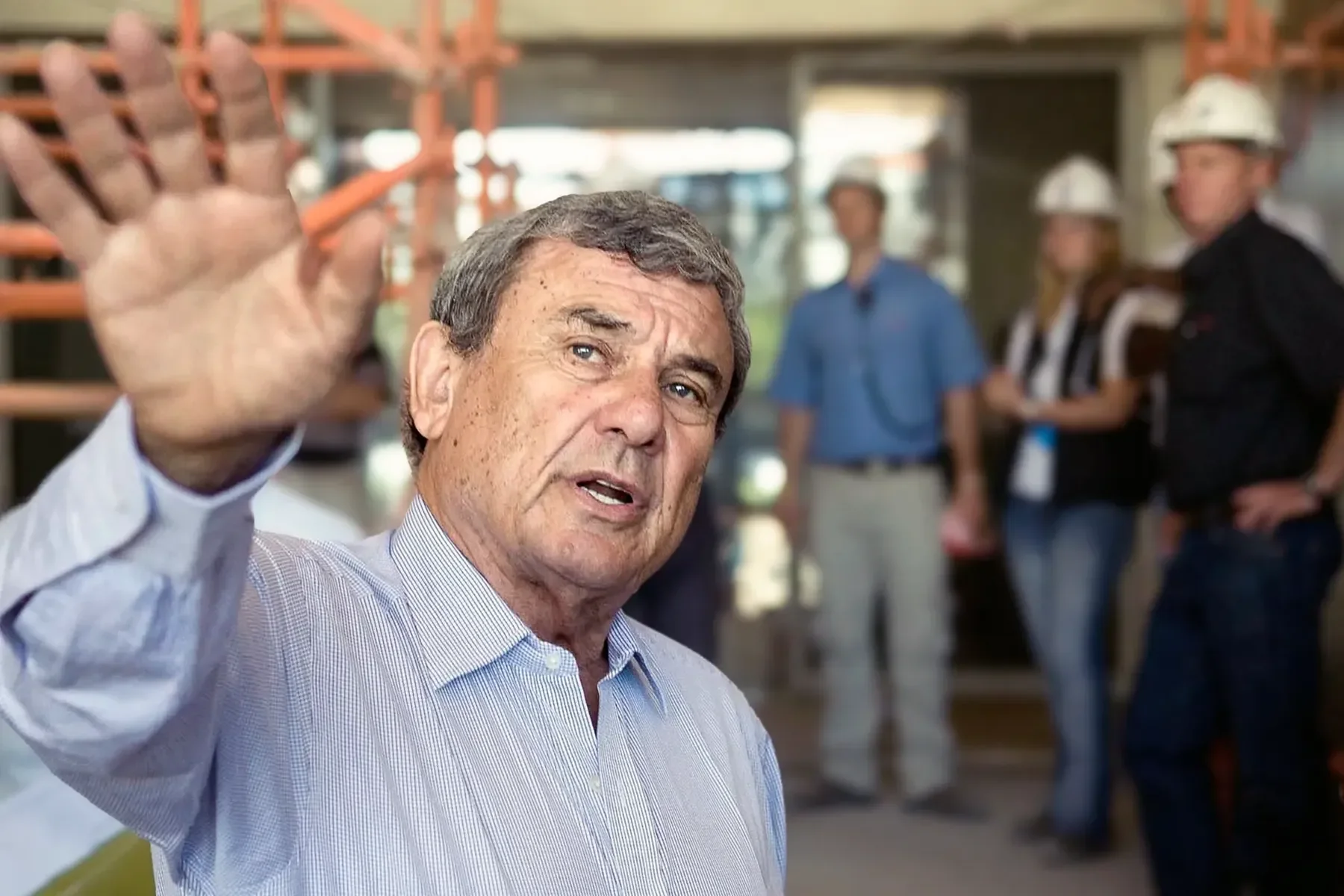 Older man with gray hair in a light blue shirt waving at the camera, with a construction site and several workers wearing hard hats in the background.