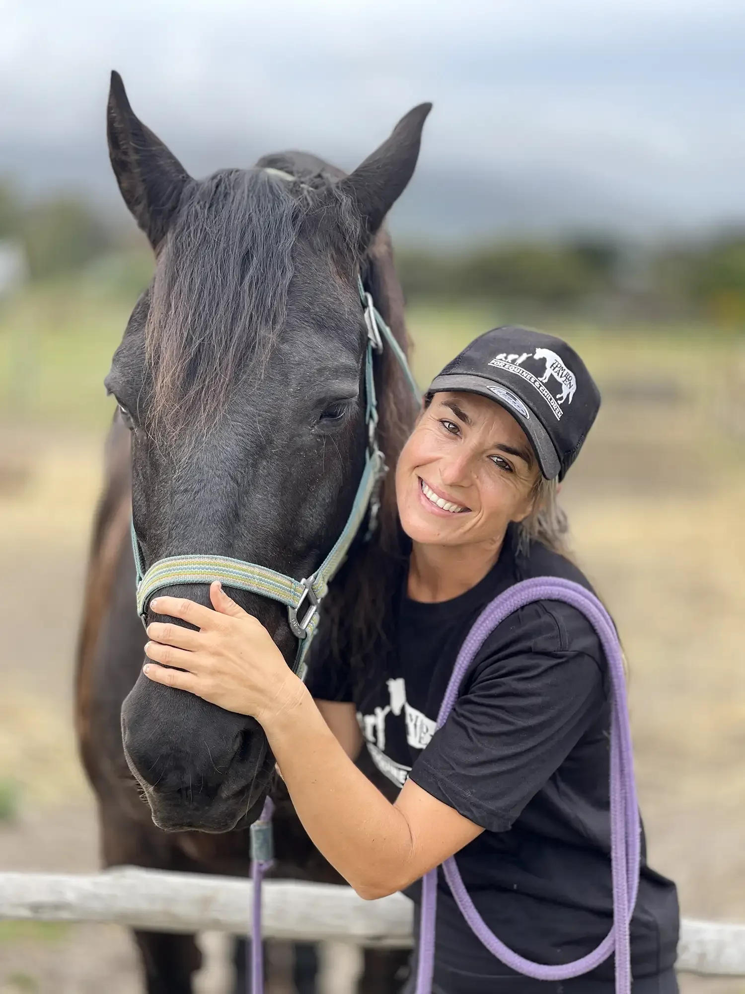 A woman hugging a black horse with a halter, smiling outdoors with a cloudy sky.