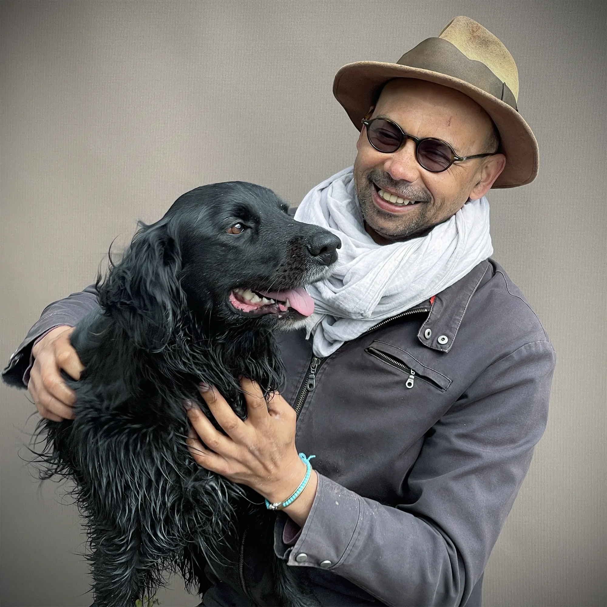 A man wearing a hat, glasses, gray jacket, and white scarf, smiling while holding a black dog with wet fur.